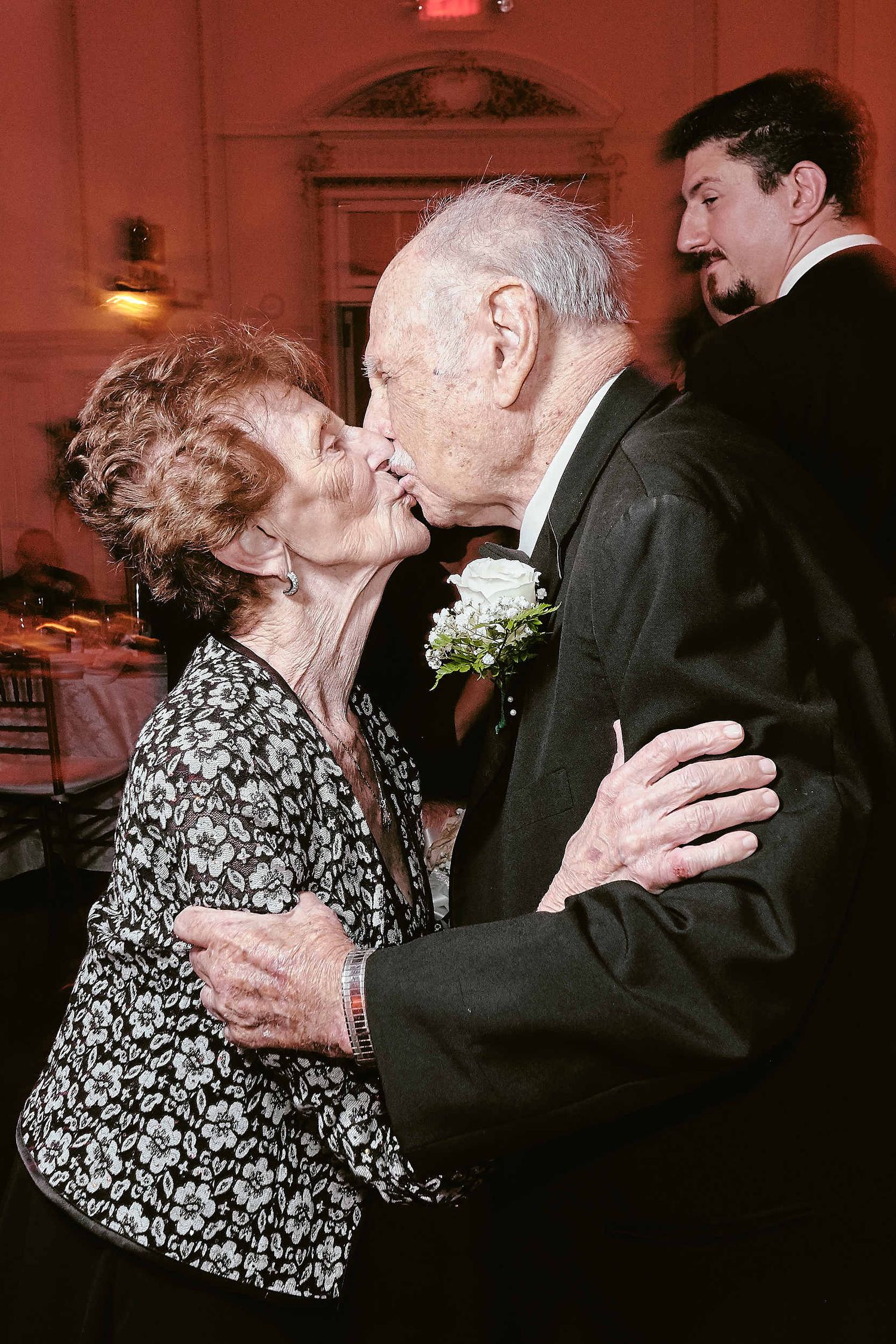 An elderly couple shares a romantic kiss while dancing at an elegant event, with a floral pattern dress and boutonniere visible.
