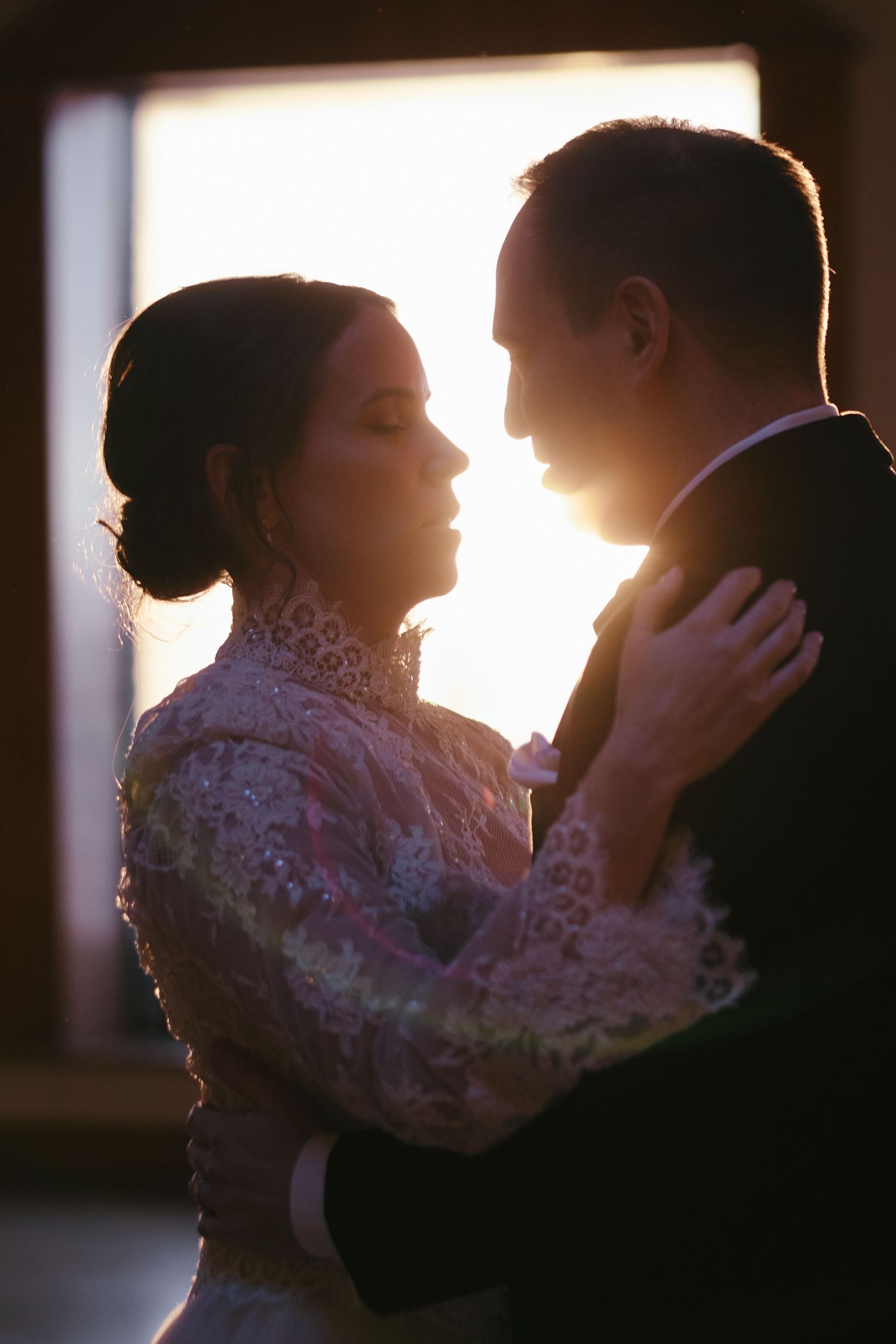 Bride and groom share a romantic backlit moment, silhouetted against golden sunset light in elegant wedding attire.