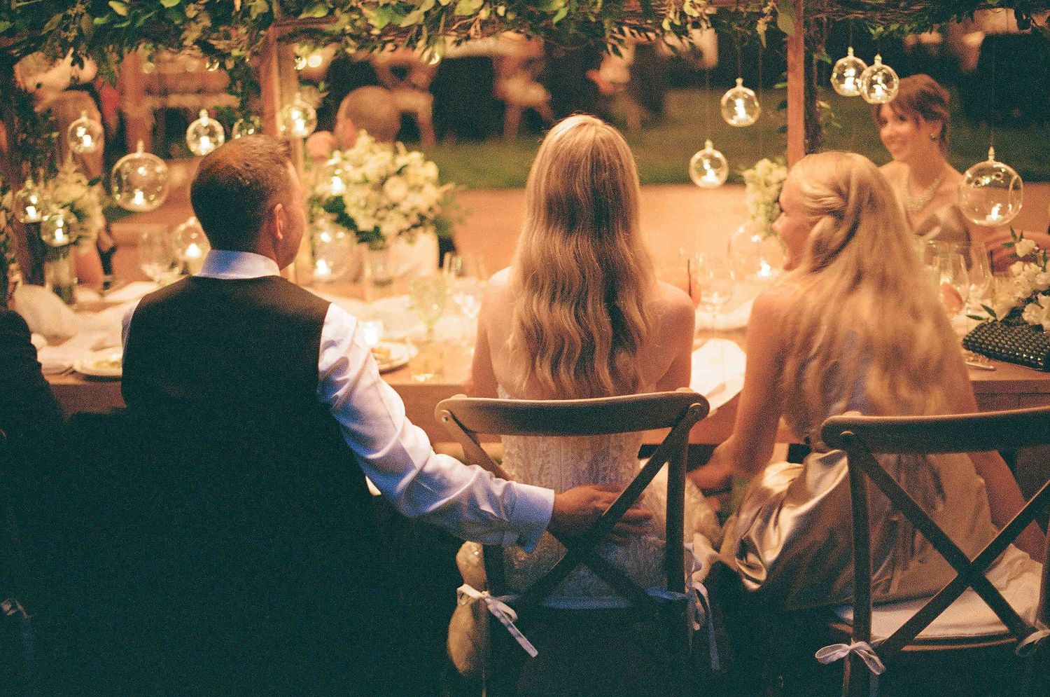 Guests seated at an elegant outdoor wedding reception with hanging lanterns and floral centerpieces at dusk.