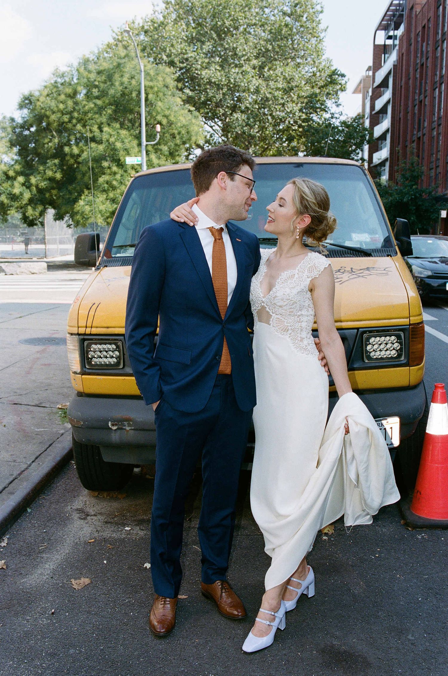 A couple in wedding attire pose together in front of a yellow vehicle on an urban street.