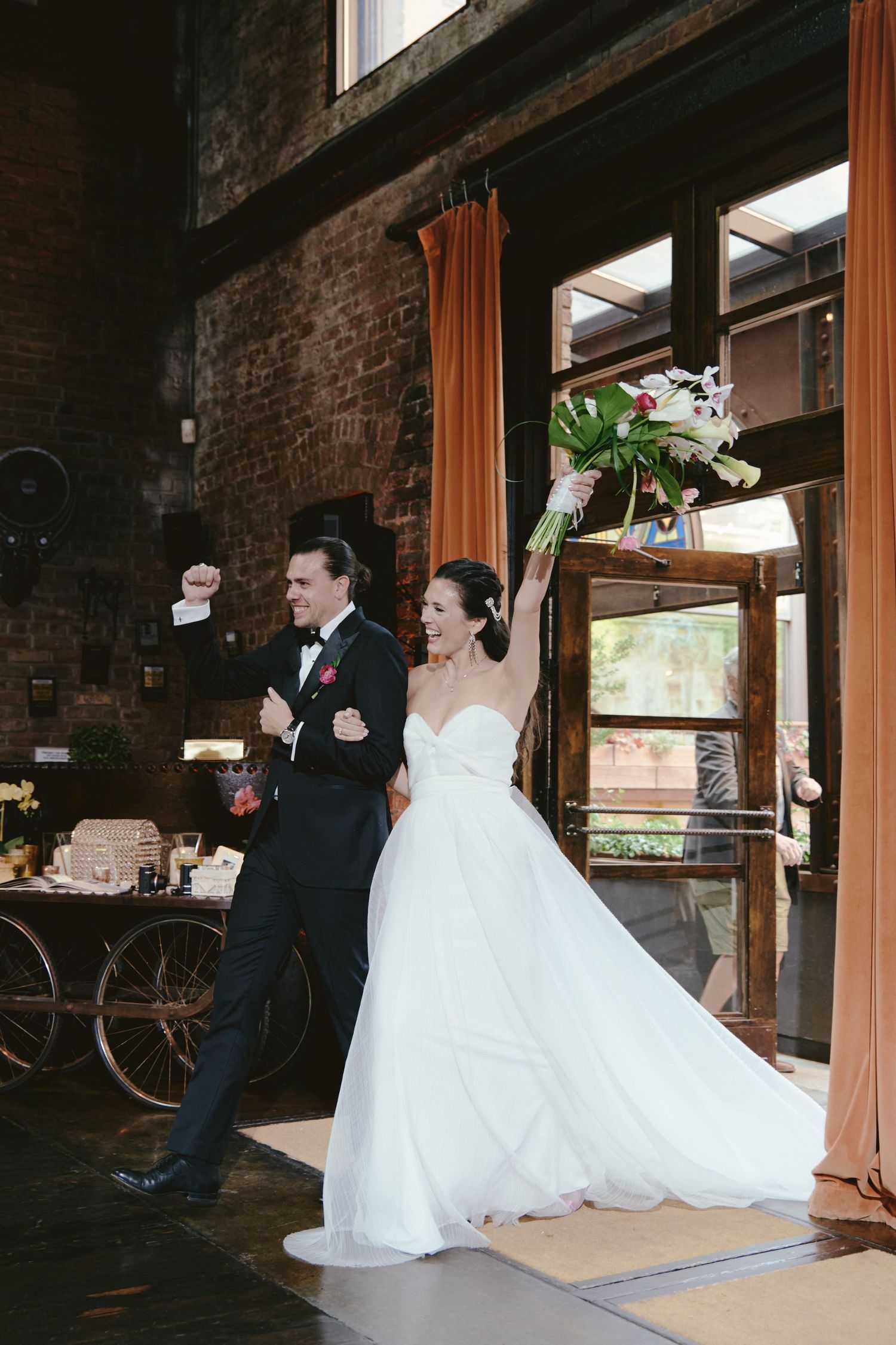 A couple celebrates joyfully entering their rustic brick wedding venue with a flowing white dress and classic tuxedo.