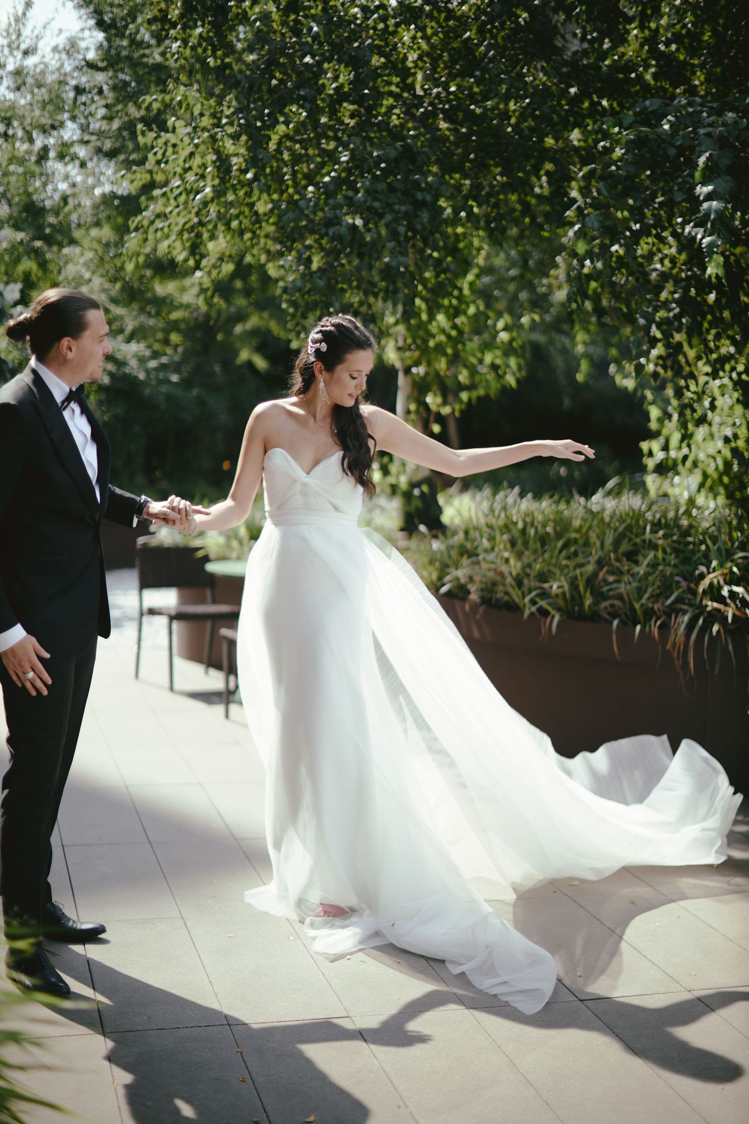 A bride in a flowing white strapless gown twirls gracefully on a sun-dappled patio while accompanied by a suited figure.