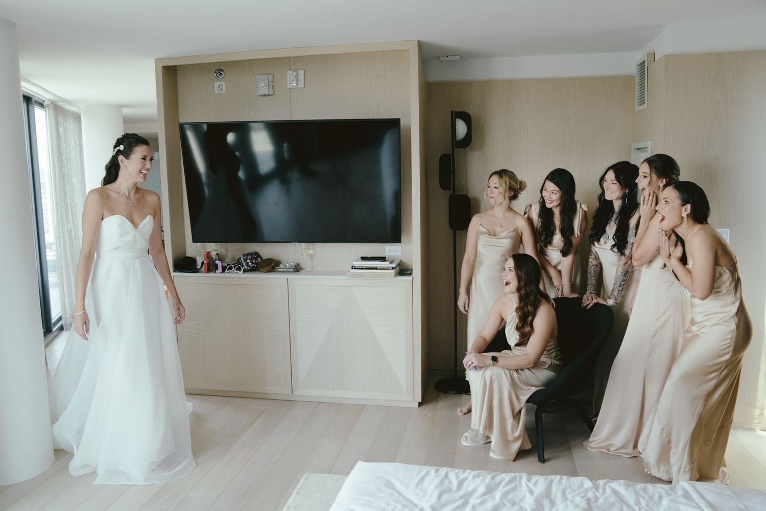 A bride in a white gown stands near a mounted TV while bridesmaids in neutral dresses gather across the room.
