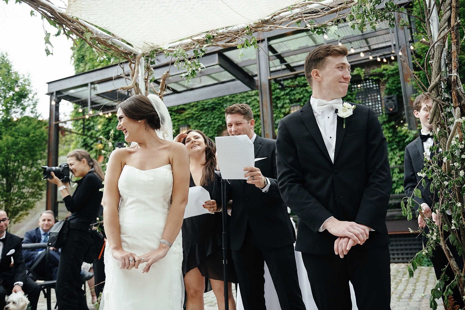A bride and groom stand at an outdoor wedding ceremony under a vine-covered pergola surrounded by guests.