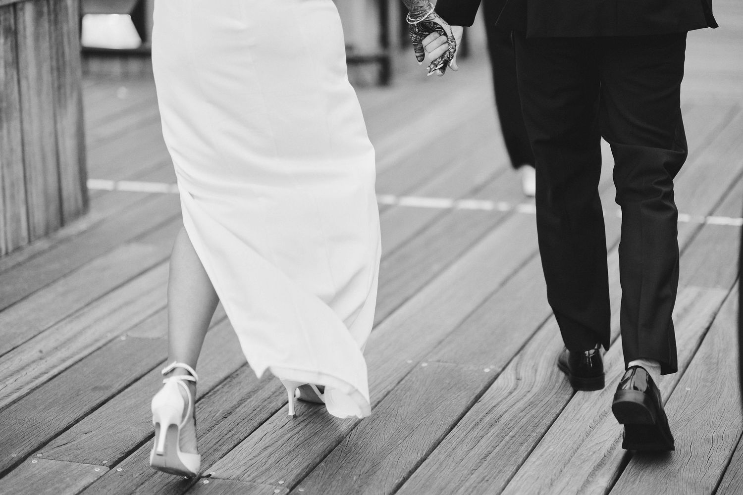 Black and white detail shot of a bride in white dress and groom in black suit walking together on a wooden deck.