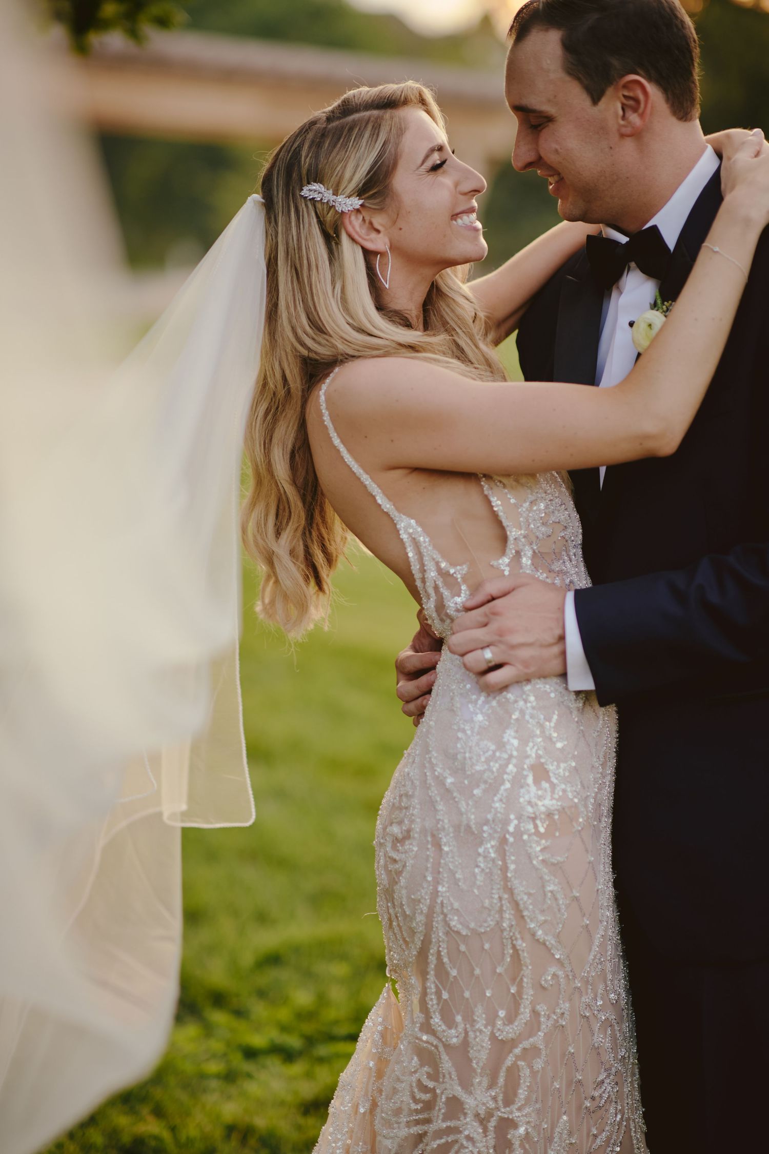 A bride in a sparkling lace gown and flowing veil shares an intimate moment with the groom in a black tuxedo outdoors.