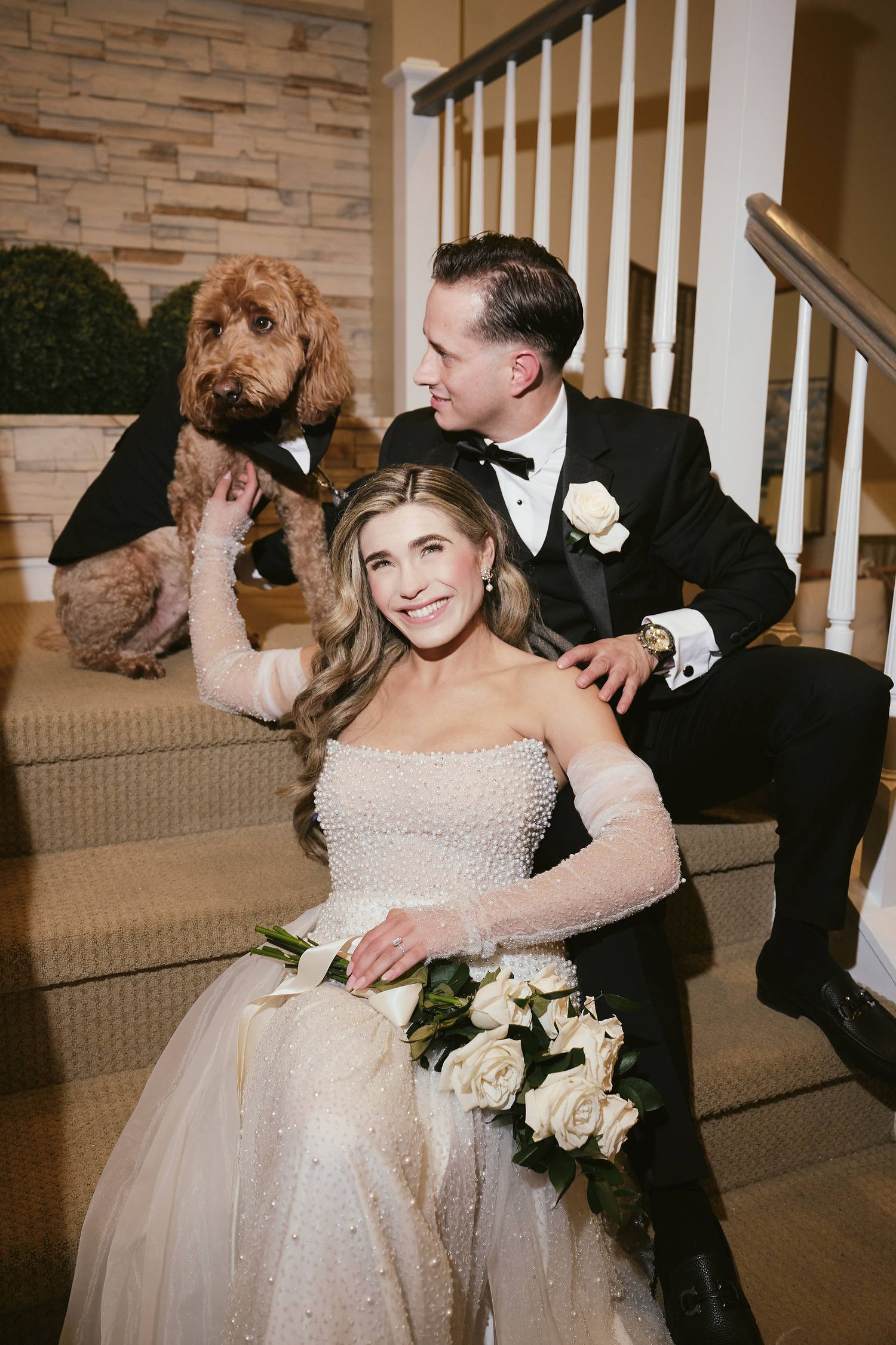 A wedding couple sits on carpeted stairs with their goldendoodle dog while holding a white rose bouquet.