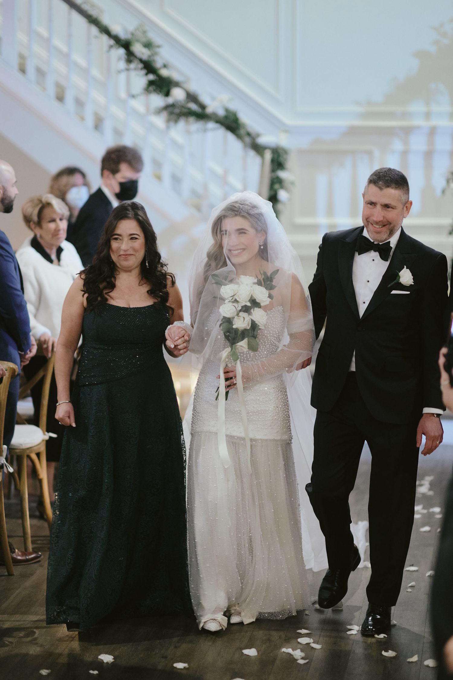 A wedding procession at an elegant venue with white walls and garland decorations along a staircase.