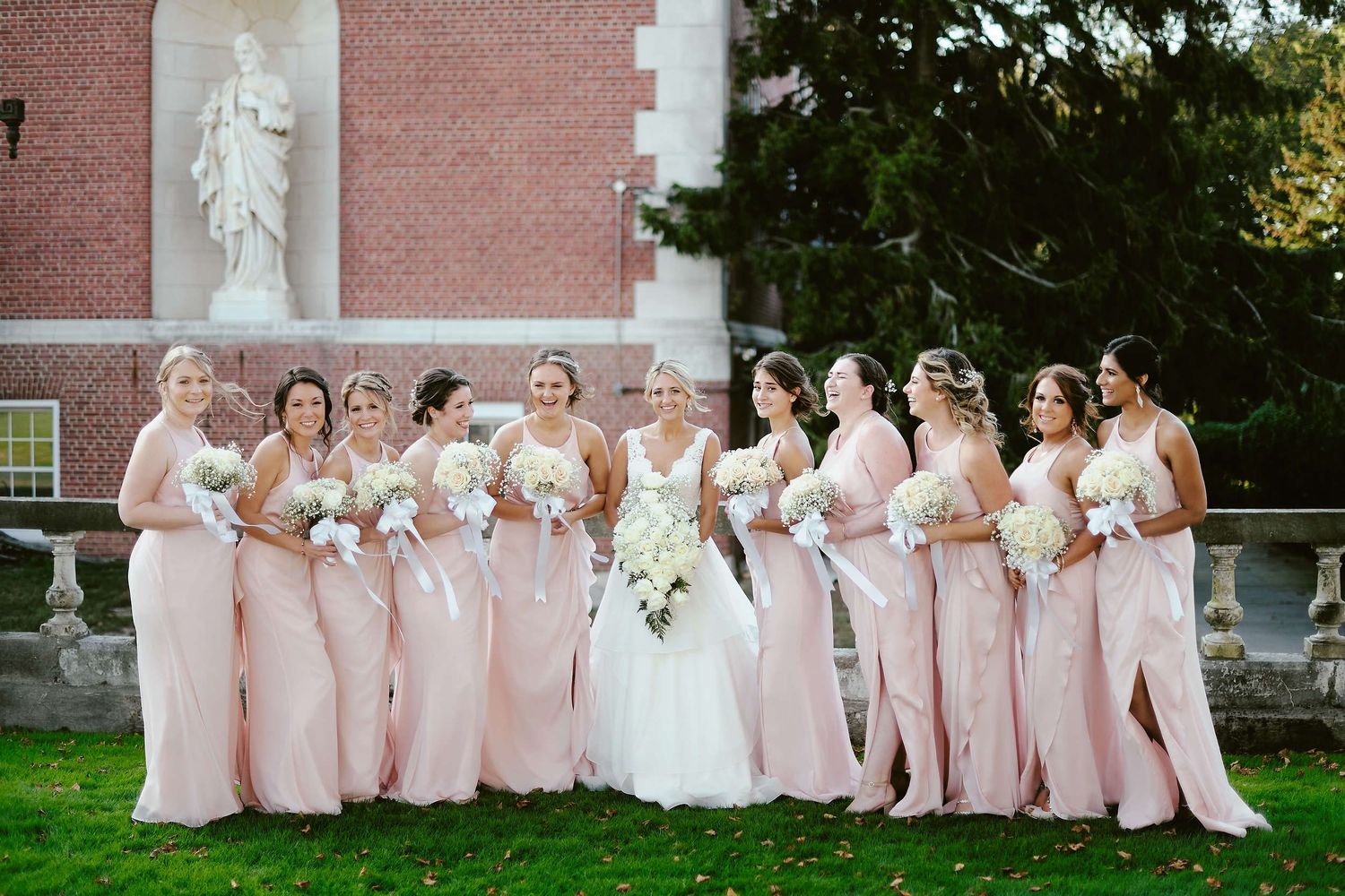 A bridal party in pink dresses poses together on a lawn in front of a brick church building with white trim.