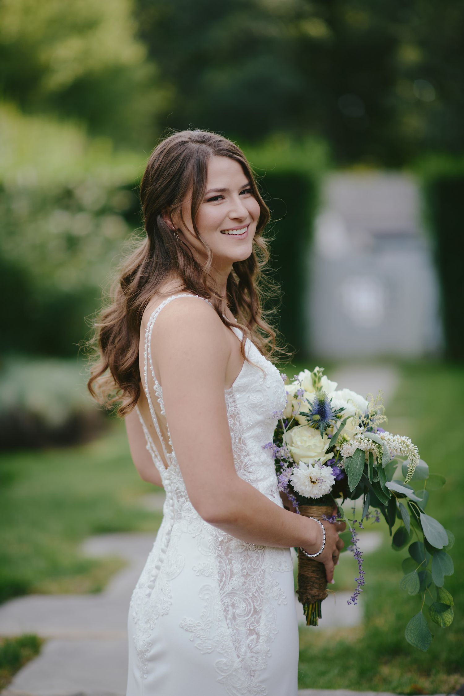 A bride in a white lace wedding dress holds a bouquet of white and blue flowers while standing in a garden setting.