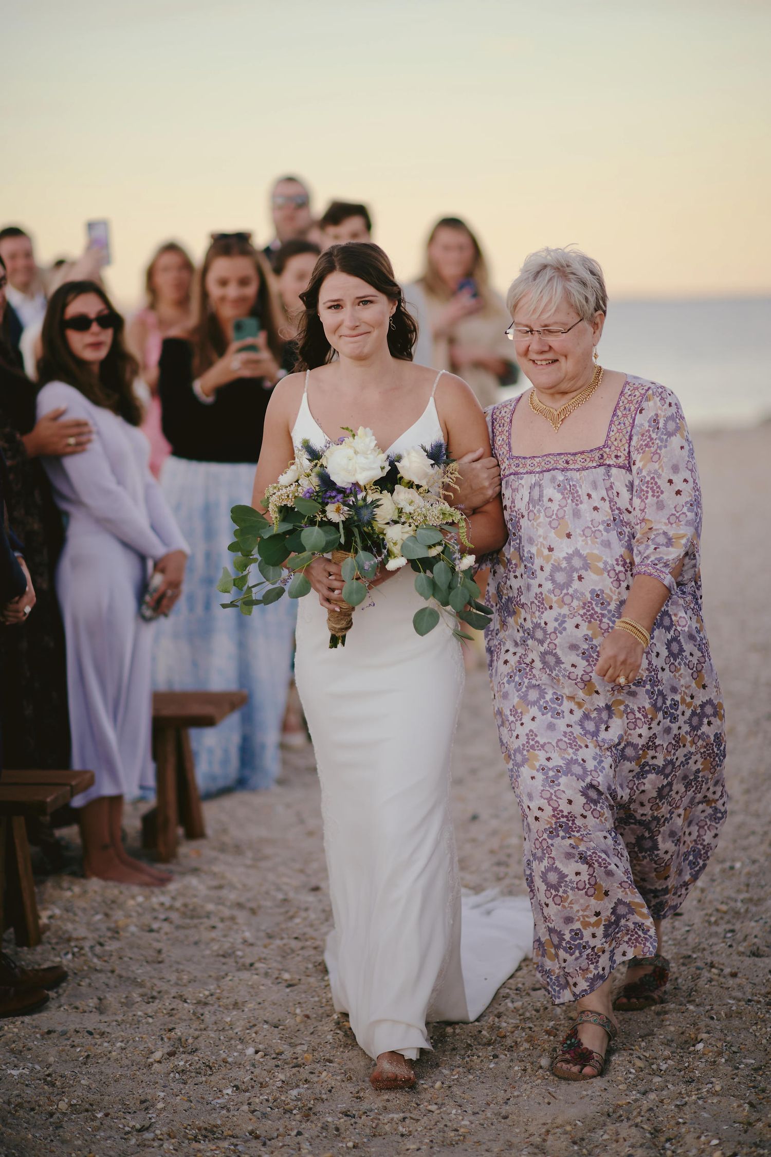 Wedding processional on sandy beach with bride carrying white bouquet accompanied by woman in floral dress.