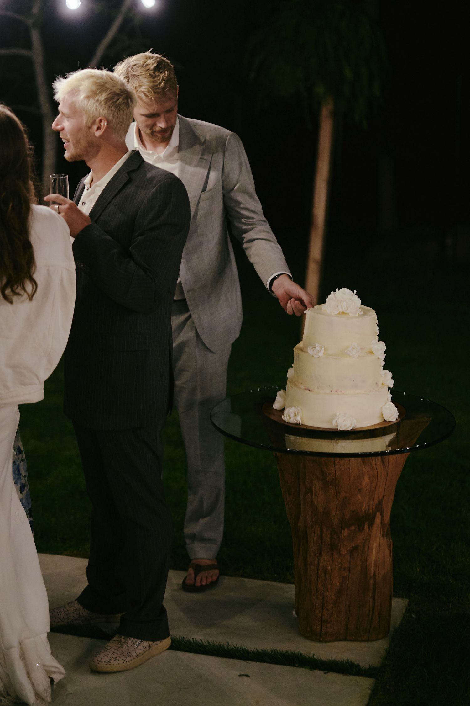 A wedding reception at night with a three-tiered white cake displayed on a rustic wooden pedestal.
