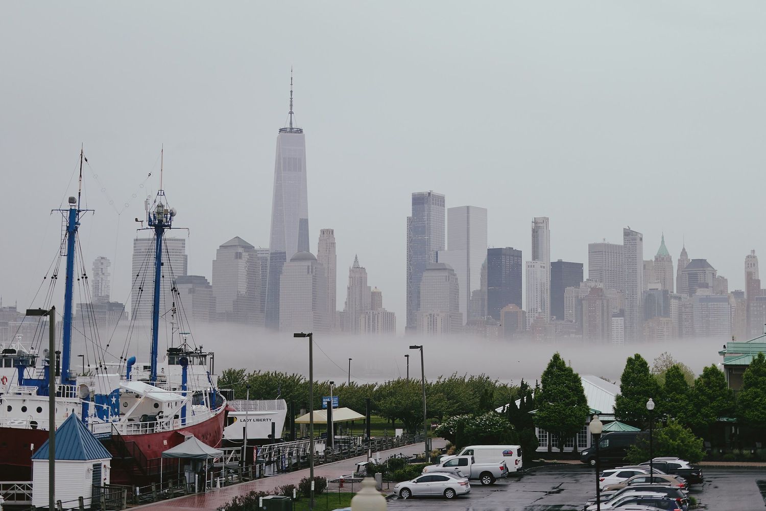 A foggy view of the Manhattan skyline with One World Trade Center visible through the mist, seen from a marina parking lot.