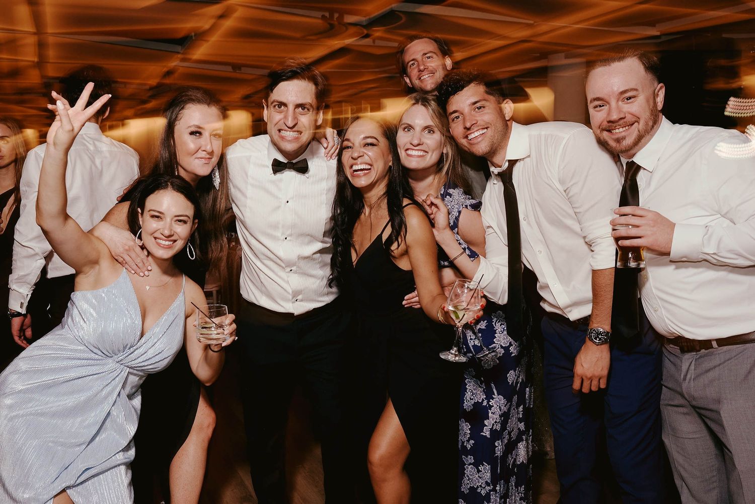 A group of formally dressed party guests pose together on a dance floor, smiling and celebrating at an indoor event.