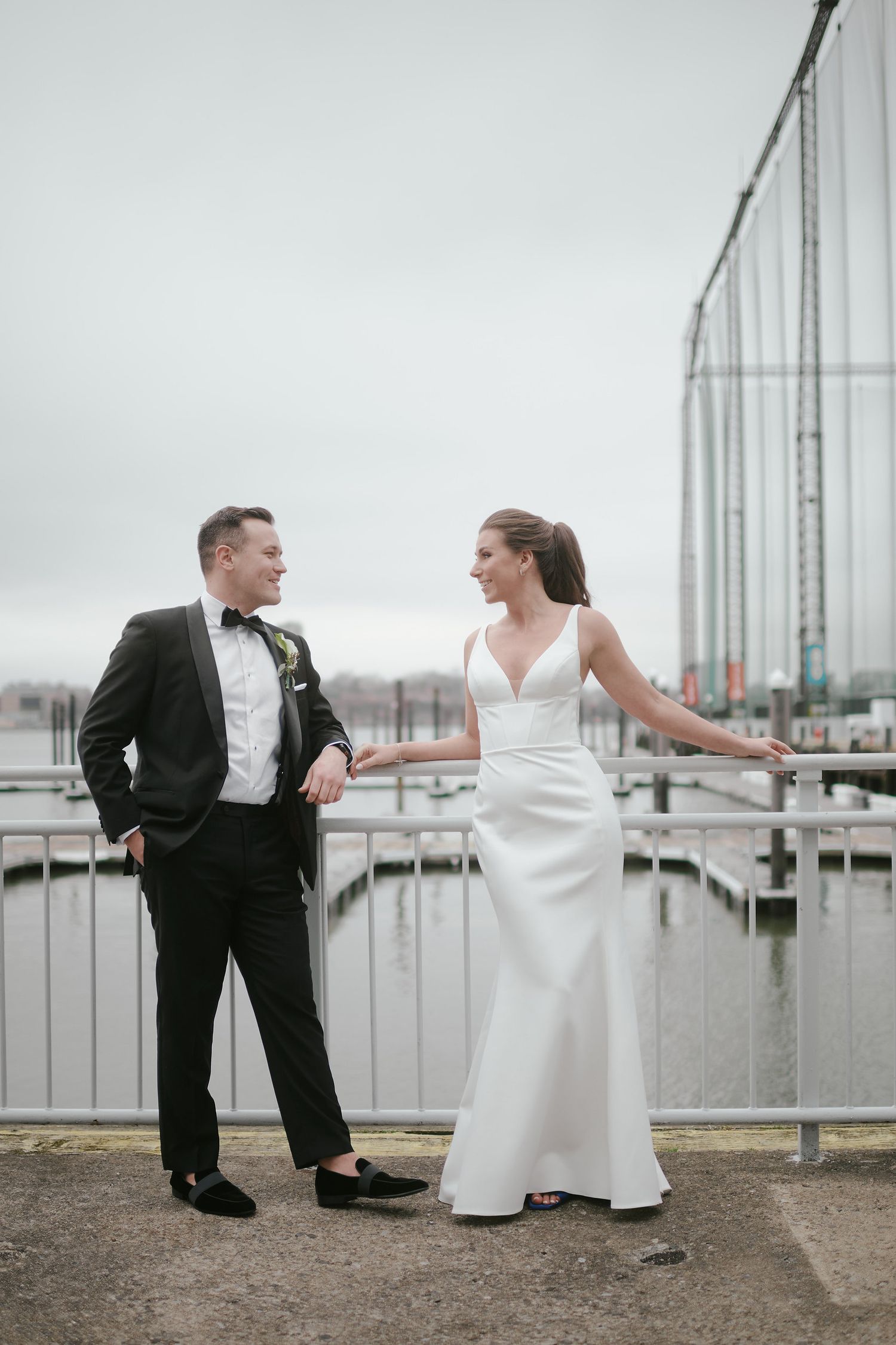 A couple in formal wedding attire share a candid moment while standing on a dock near glass railings and marina in overcast weather.