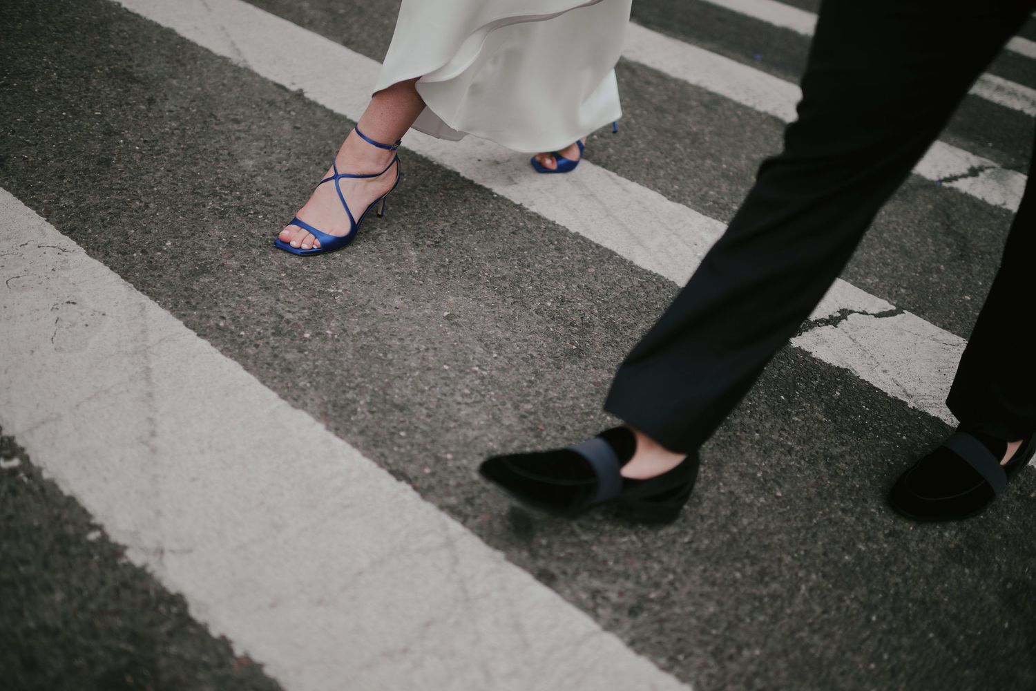 Two pairs of feet in contrasting footwear stand at a crosswalk, one in blue sandals and one in black flats on asphalt.