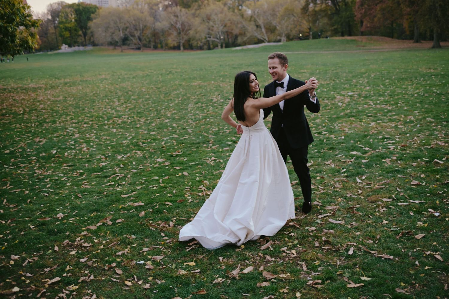 A wedding couple dances together on a grassy lawn with fallen autumn leaves.