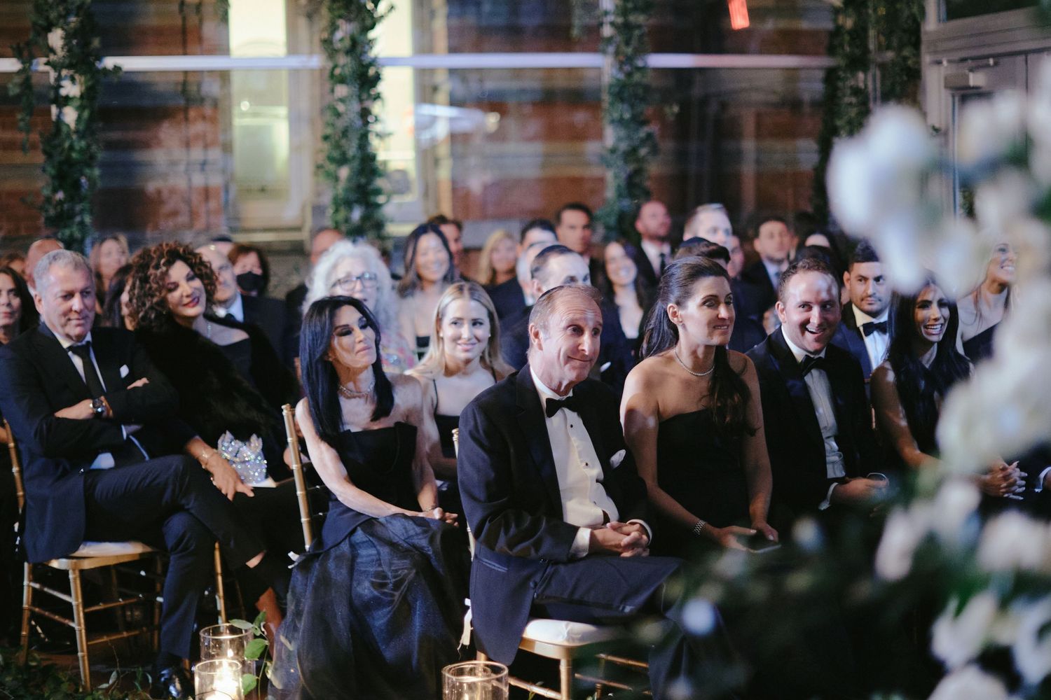 Wedding guests sit in elegant formal attire watching a ceremony in an intimate indoor venue decorated with greenery.