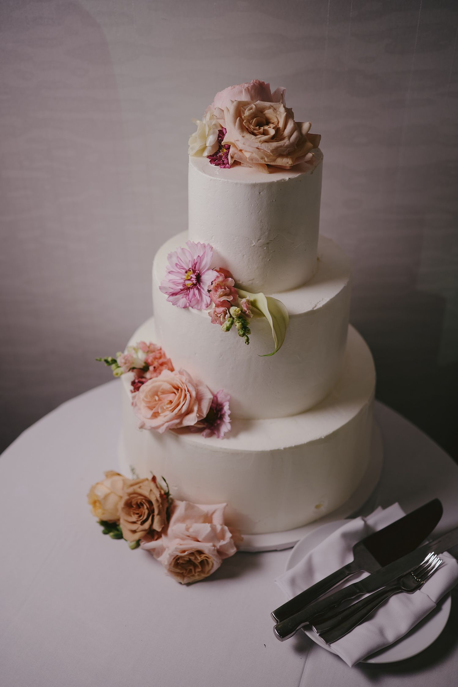 Elegant three-tier white wedding cake decorated with soft pink and blush roses on display with serving utensils.