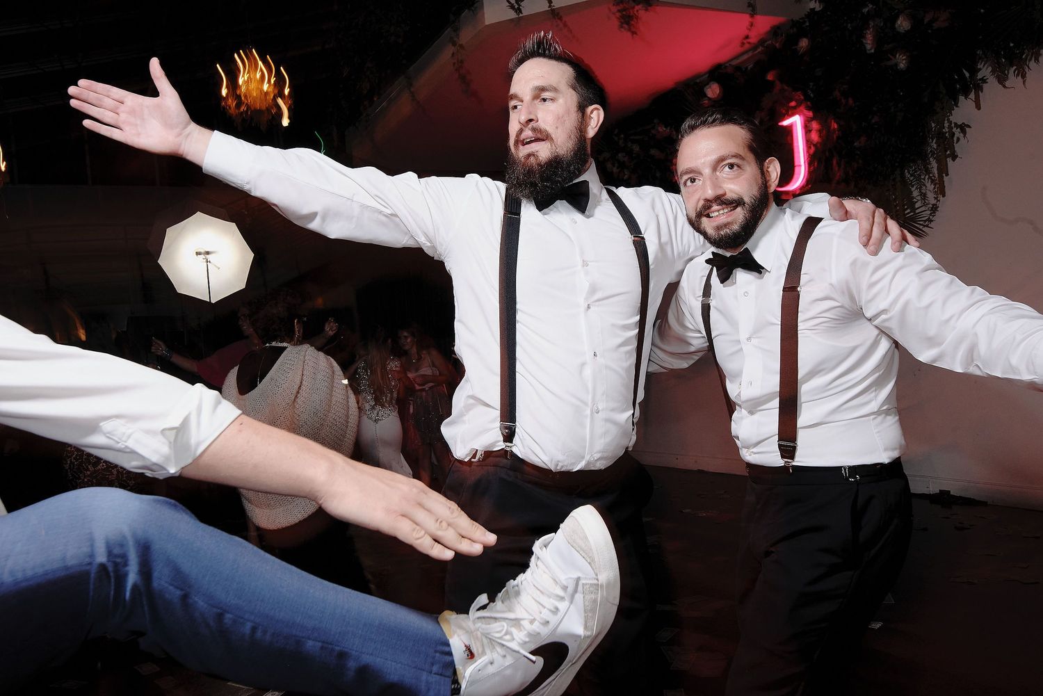 Partygoers in suspenders enthusiastically dancing at a celebration while someone jumps in foreground.