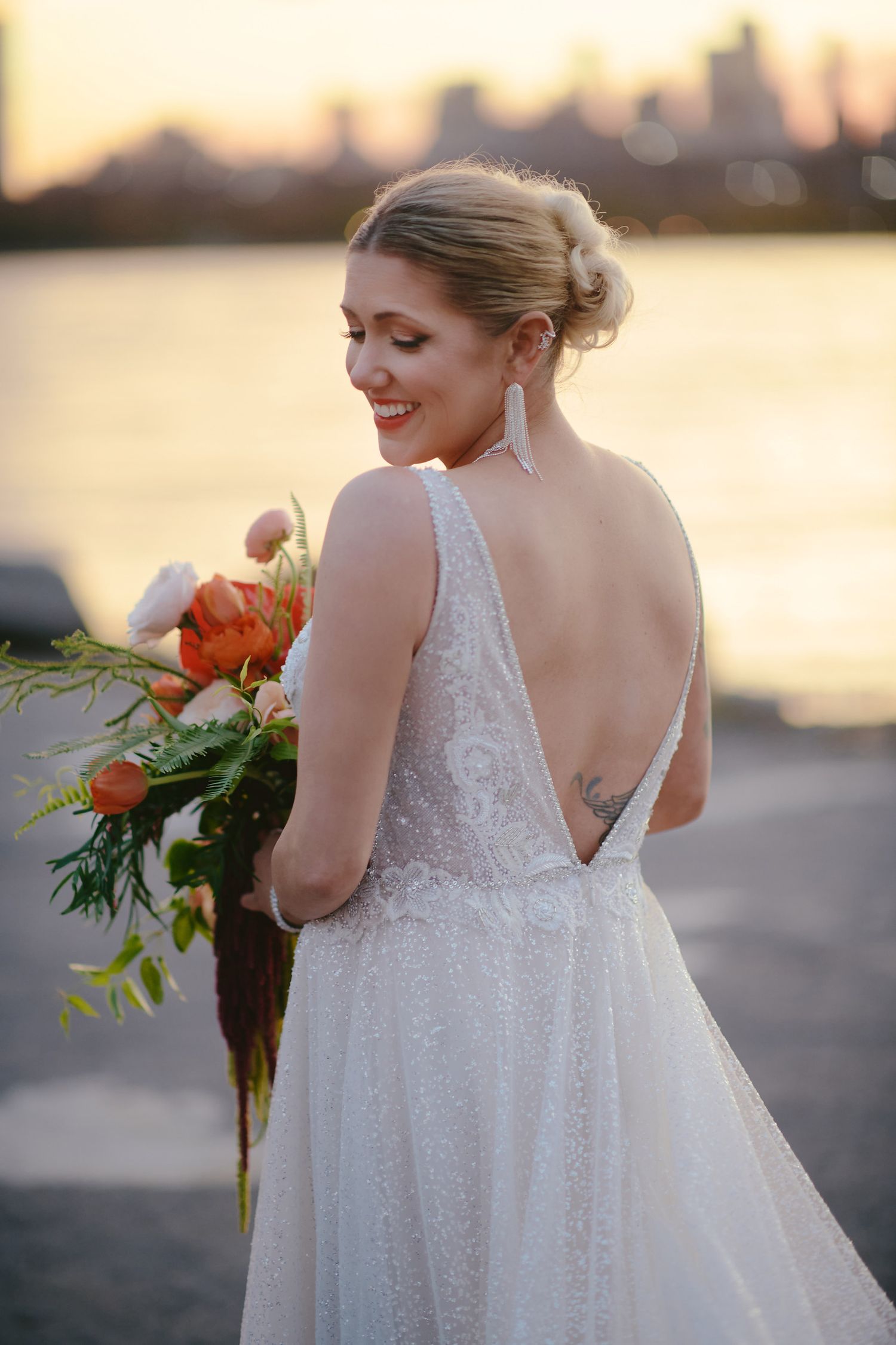 A bride in a white lace v-back wedding dress holds a vibrant orange bouquet at sunset by the water.