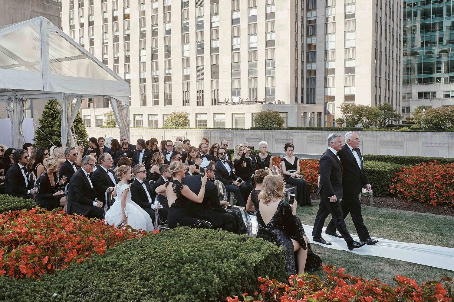 Wedding guests gather at an outdoor ceremony with modern architecture and vibrant orange flowers in the landscaping.