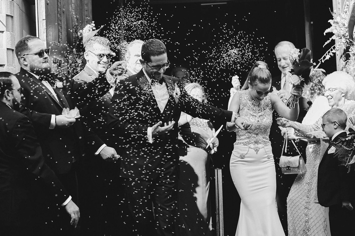 Wedding celebration captured in black and white as guests shower the newlyweds with confetti outside a venue.