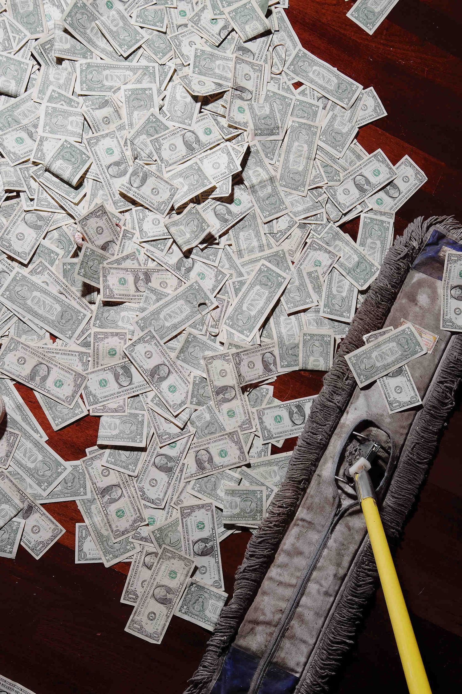 A pile of US dollar bills scattered across a dark wooden surface with a broom or sweeping tool visible on the side.