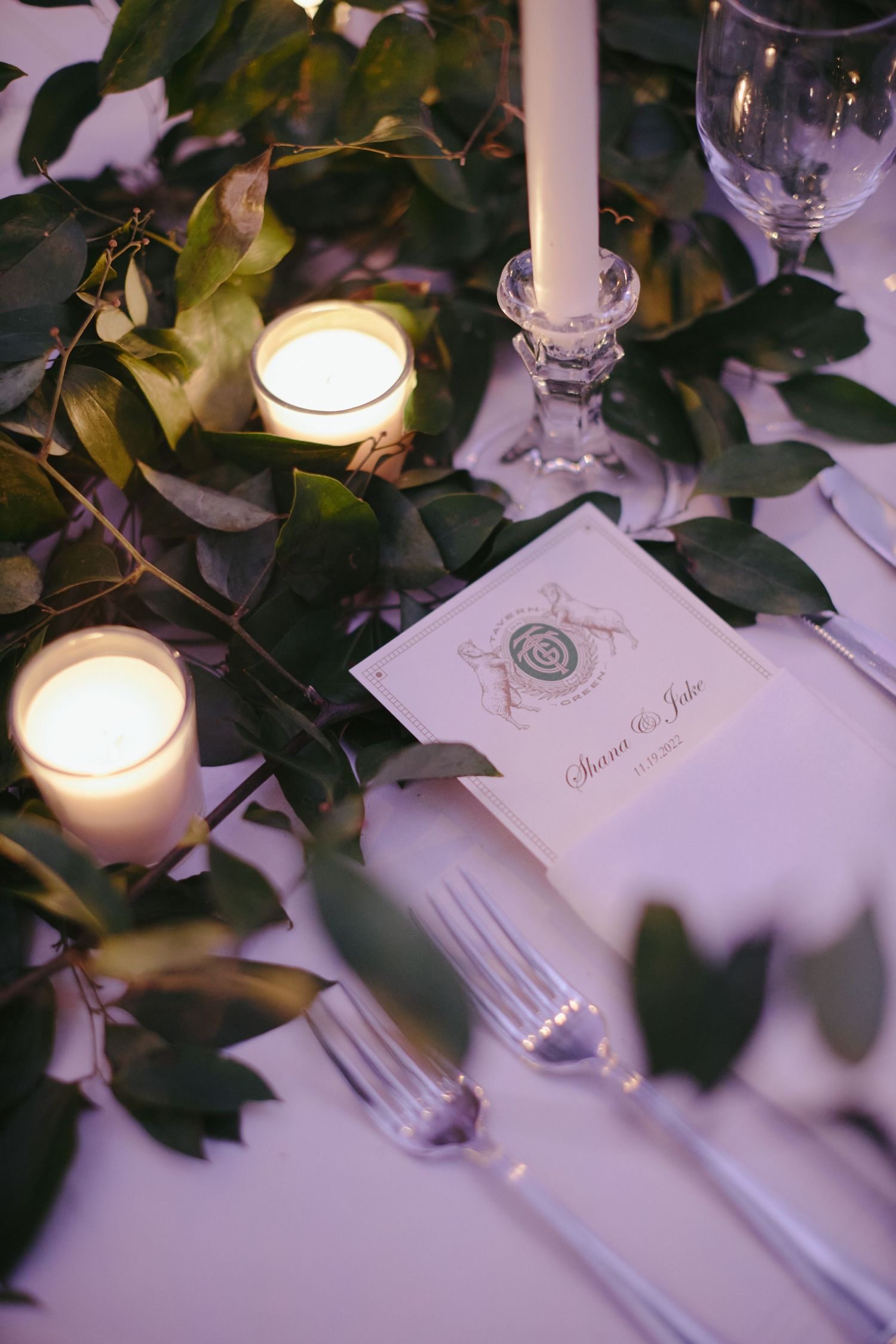 An elegant place setting with lit candles, greenery, and a white invitation card on a crisp tablecloth.