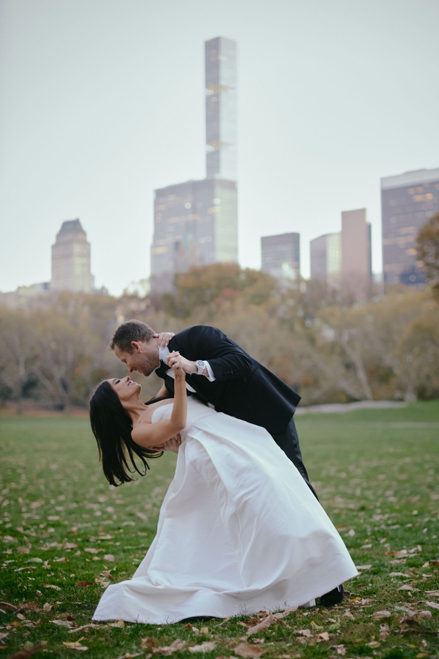 A romantic dip kiss between newlyweds in Central Park with Manhattan skyline and autumn foliage in the background.