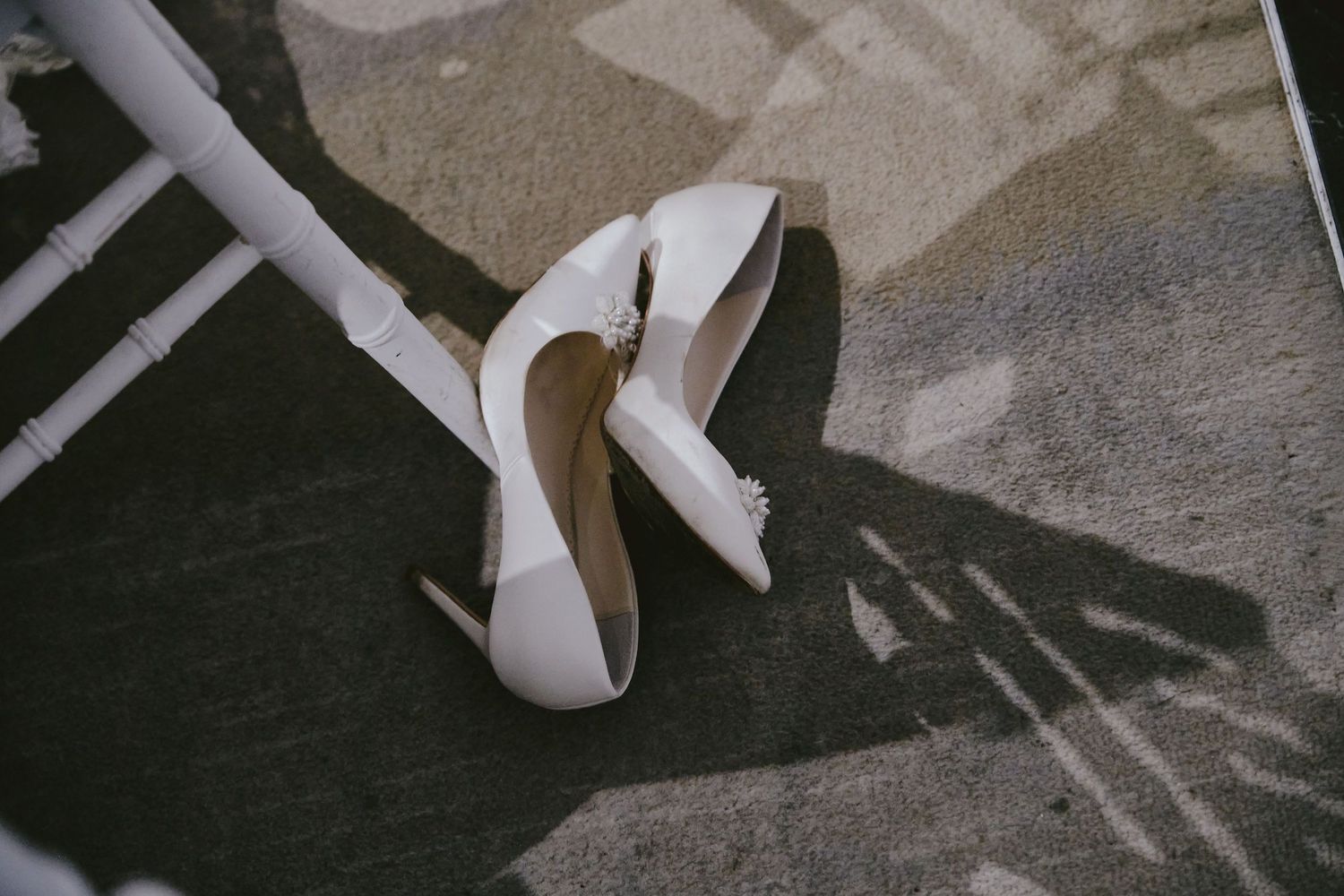 A pair of elegant white high heel shoes on a gray carpet casting long shadows.