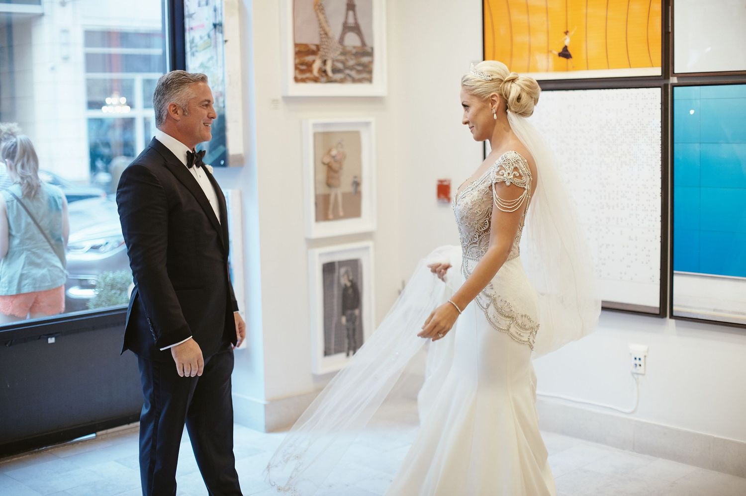 First look between a bride in a fitted lace gown and groom in a black tuxedo at an art gallery venue.