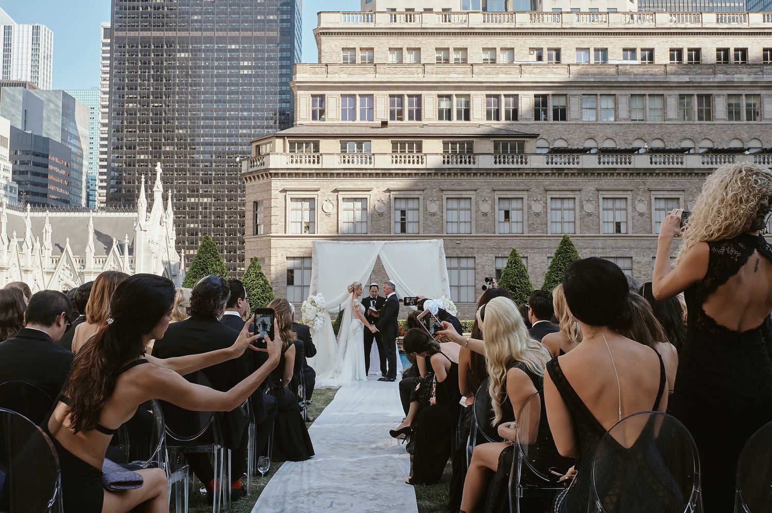 Guests capture photos with phones during an outdoor wedding ceremony on a city rooftop with cathedral backdrop.