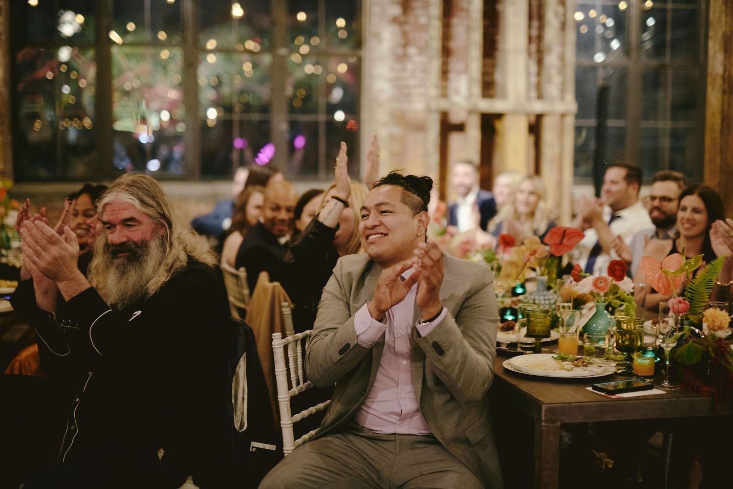 Wedding reception guests applaud during an indoor celebration at a rustic venue with exposed brick walls and floral centerpieces.