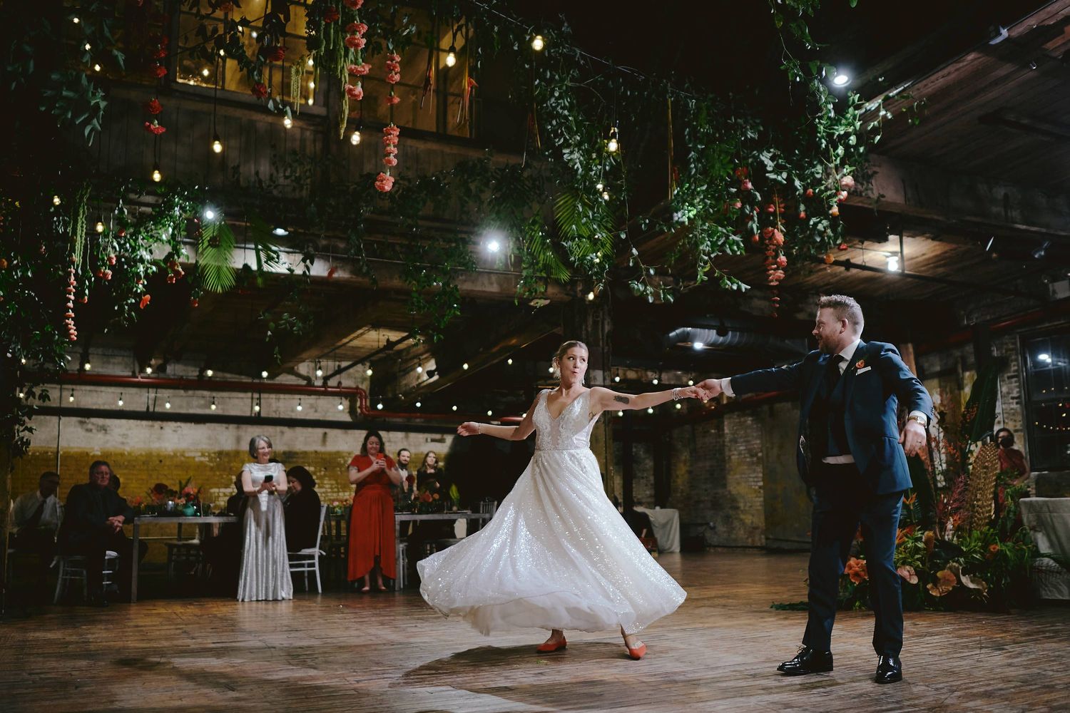 A couple dances together at an evening reception in a rustic venue decorated with twinkling lights and greenery.