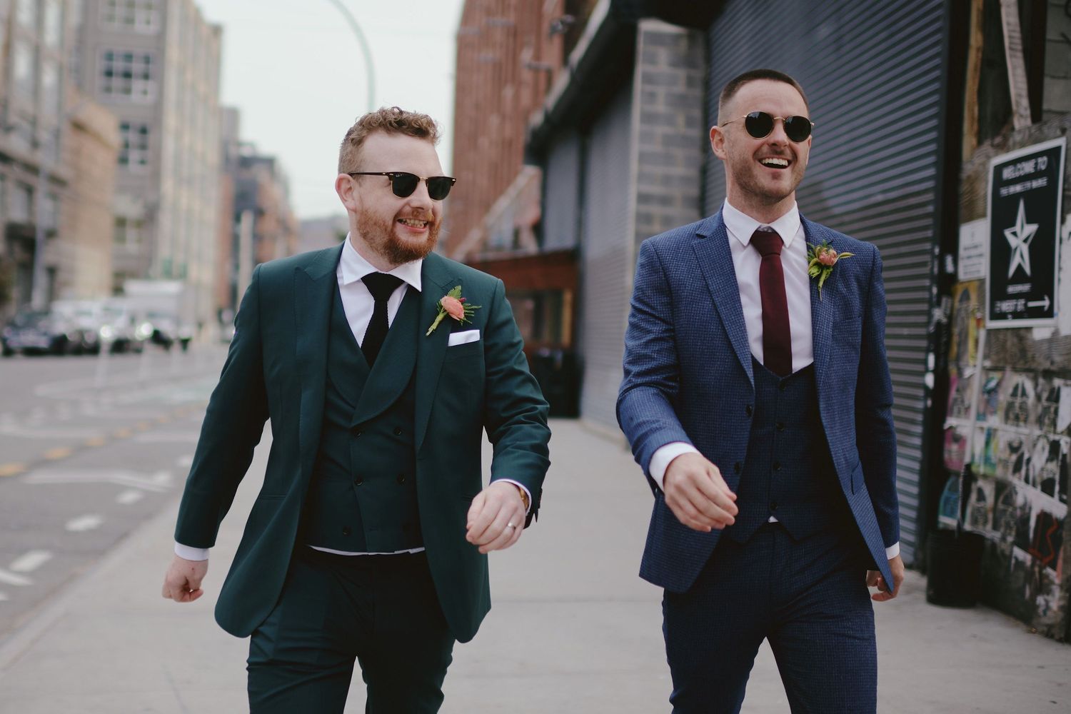 Two men in stylish suits walking down an urban street, wearing sunglasses and looking confident.