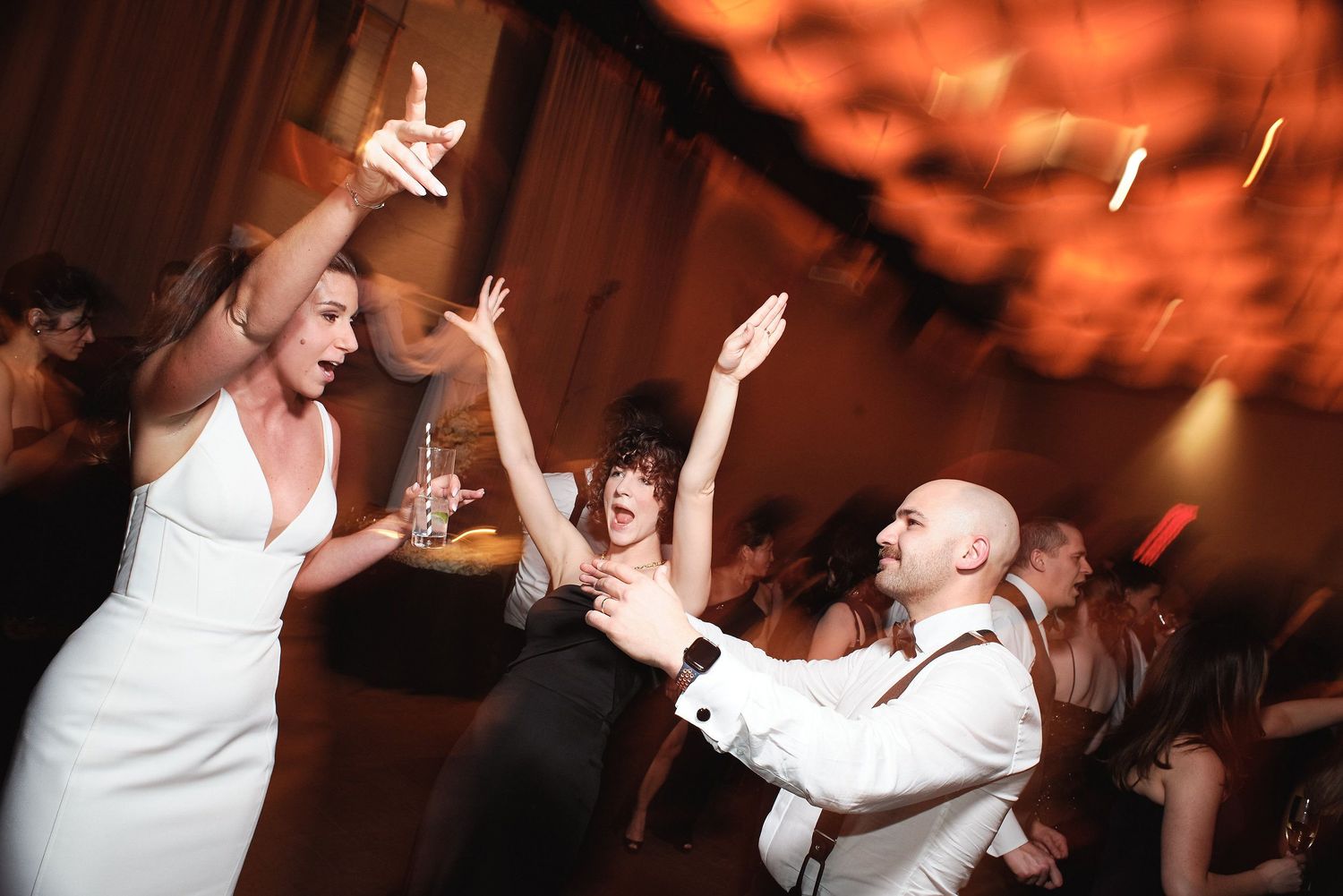 People dancing and celebrating with raised arms at a wedding reception with orange ambient lighting in the background.