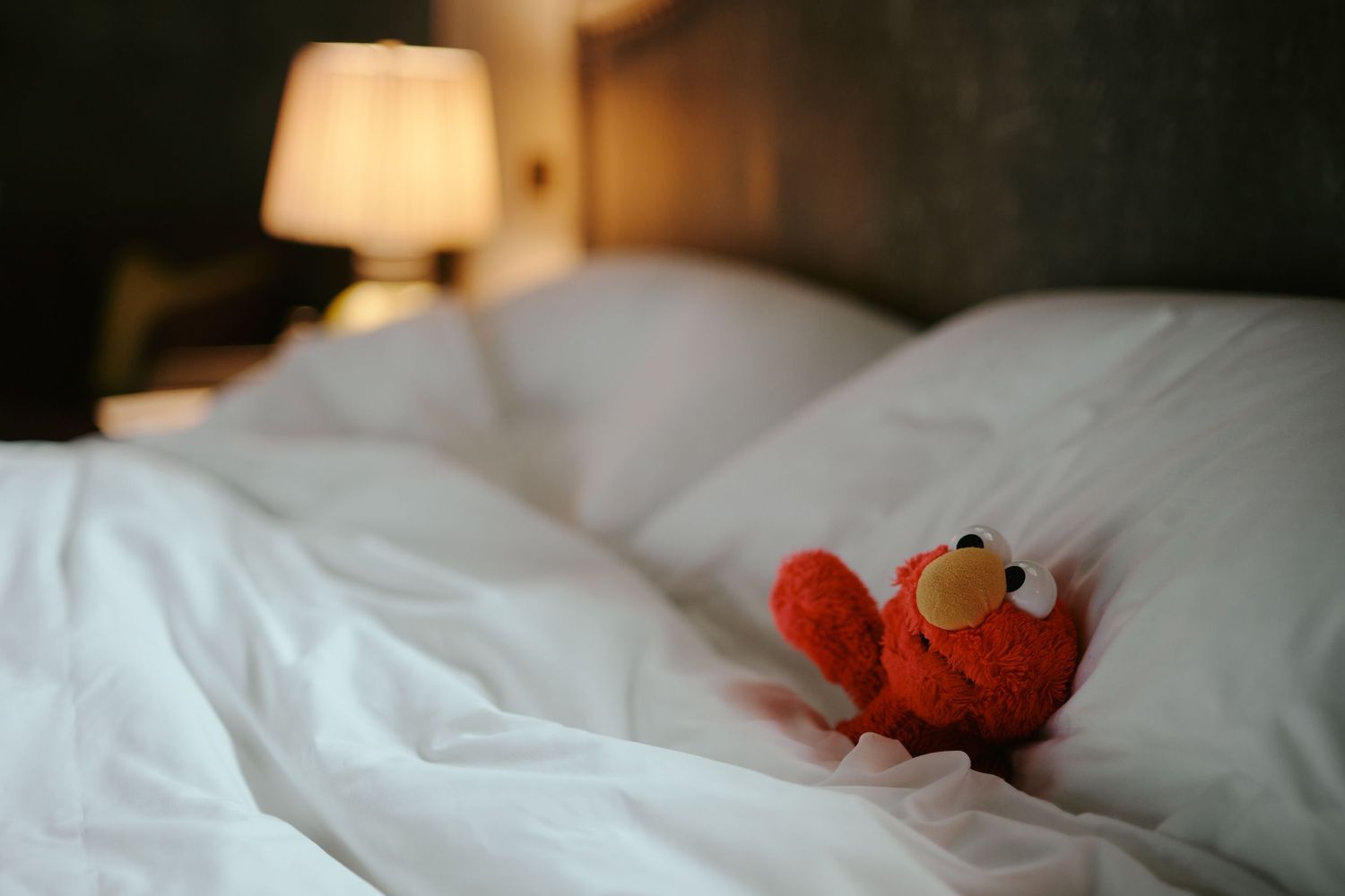A small Elmo plush toy rests on white rumpled bedsheets with a soft lamp glowing in the background.