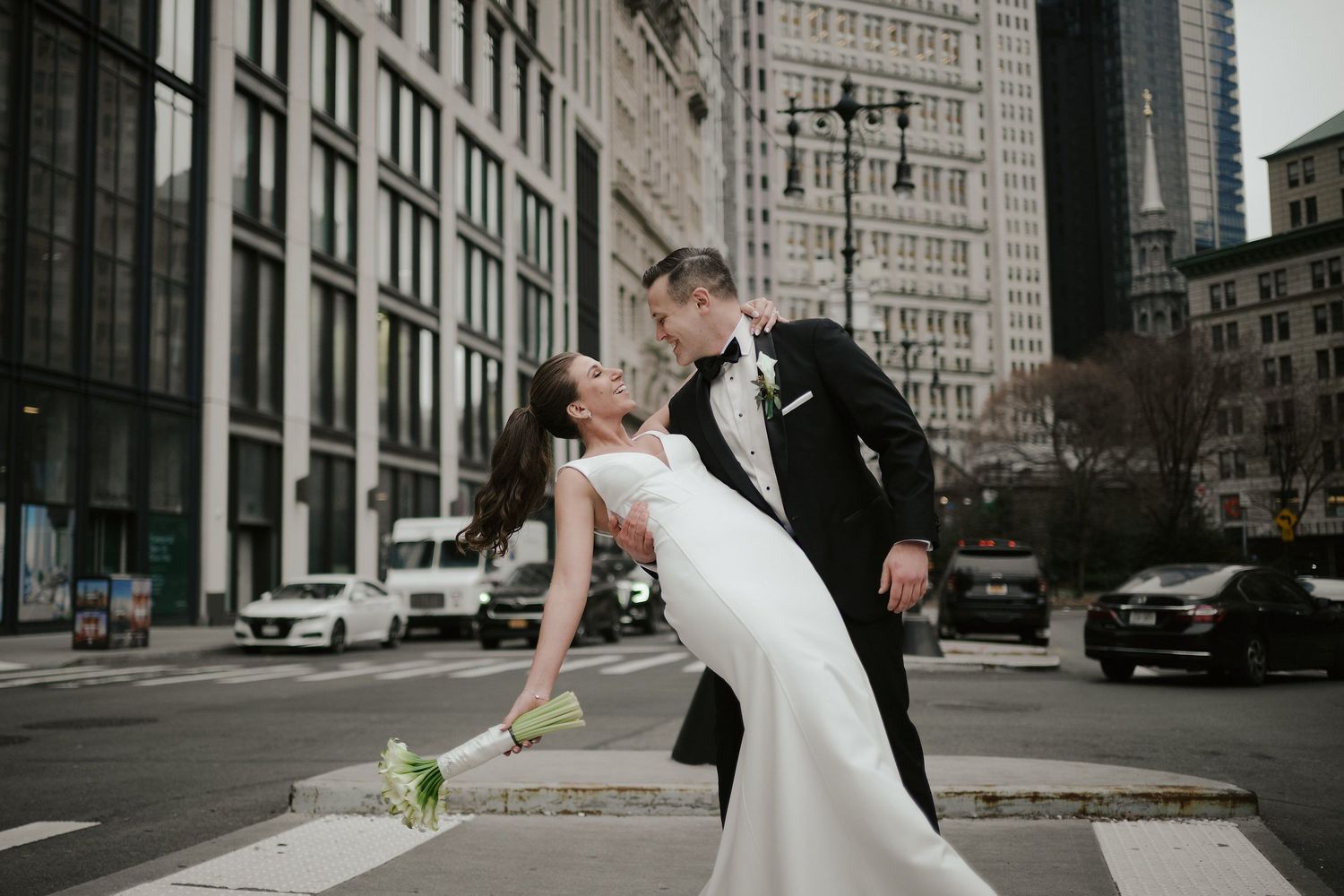 Romantic wedding couple in elegant attire dips on city street with tall buildings and traffic in background.