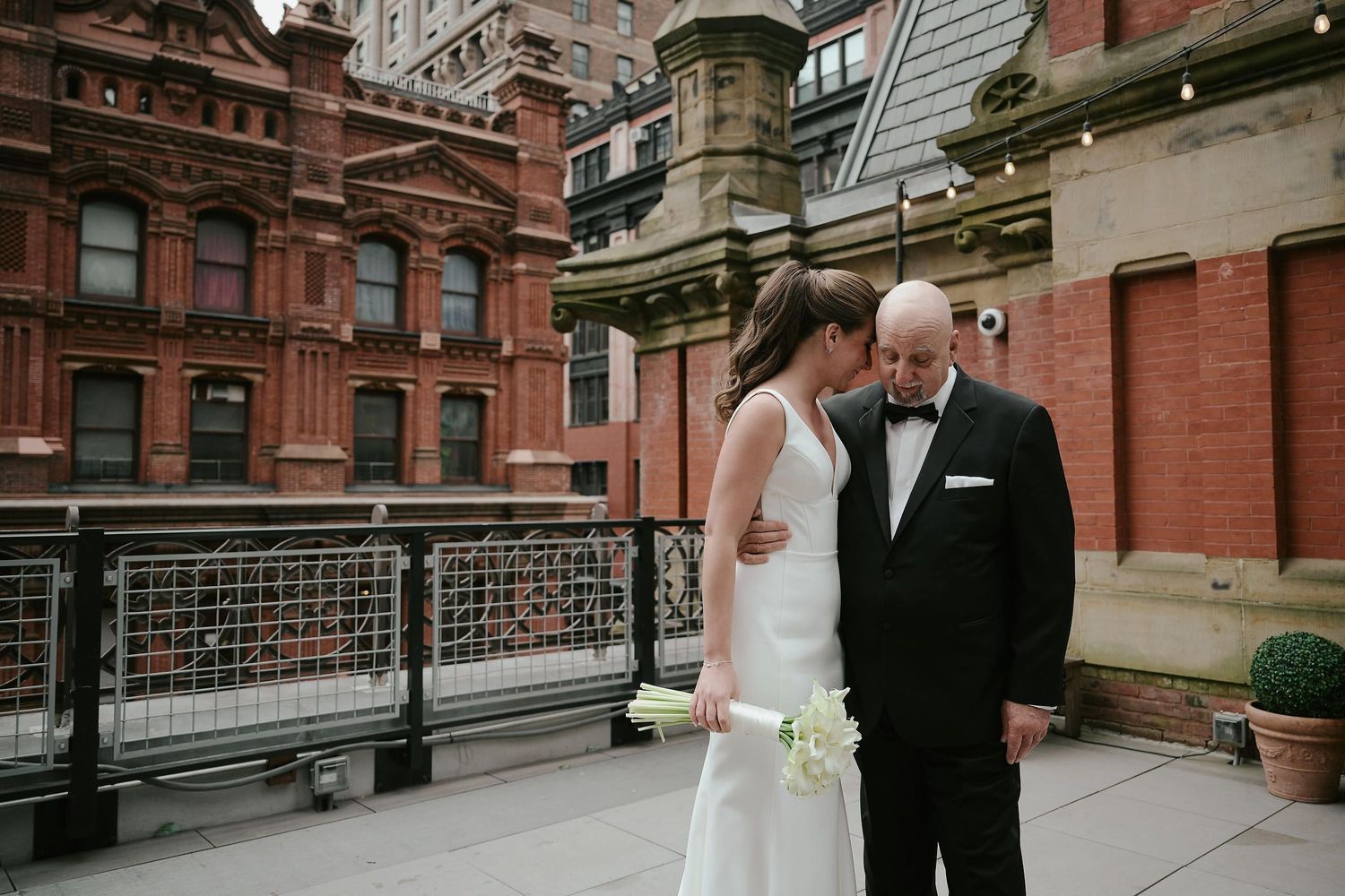A couple in wedding attire sharing an intimate moment on a rooftop with brick buildings in background.