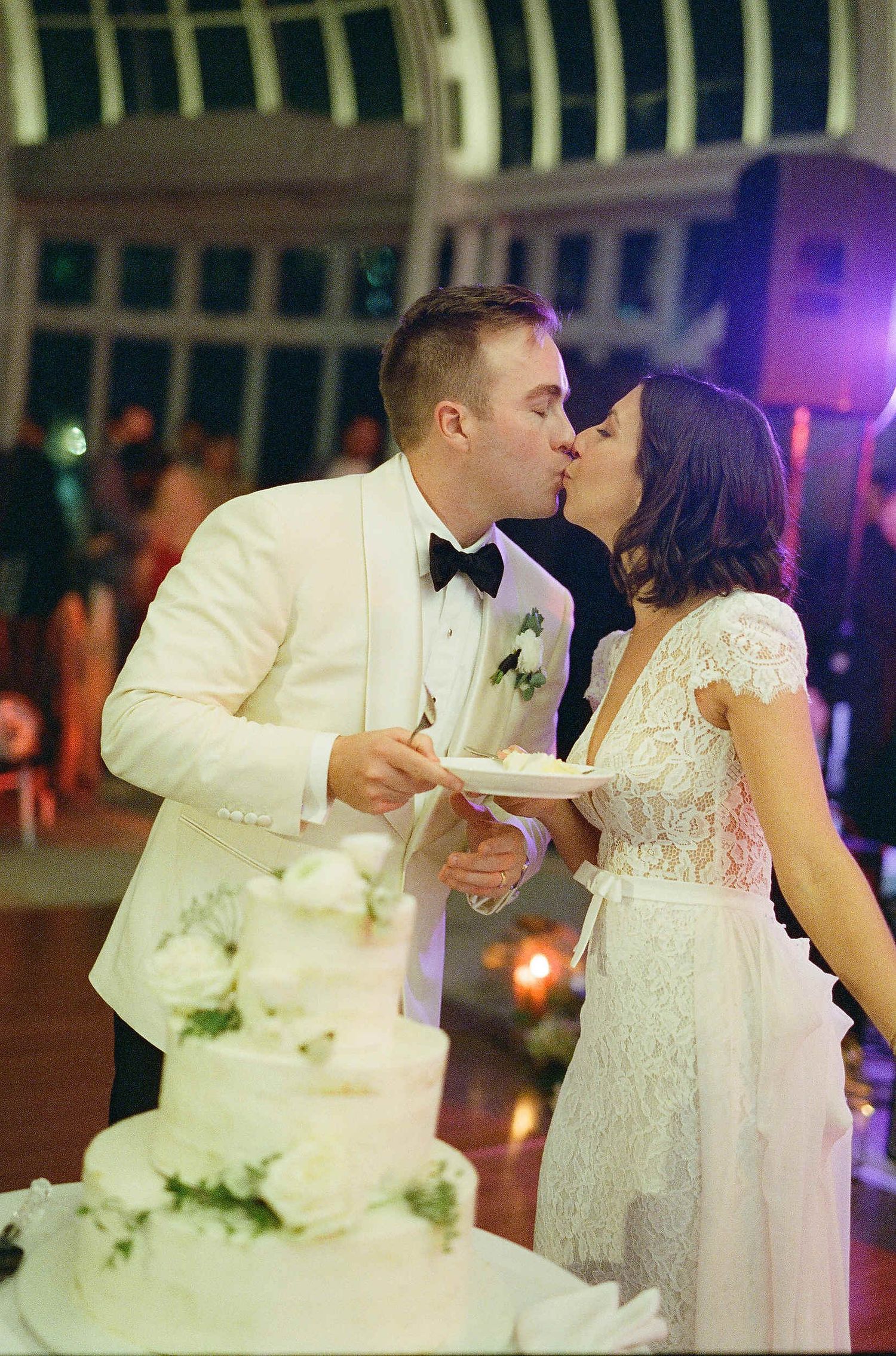 A couple in formal wedding attire sharing cake and kissing at their reception with a white tiered cake in the foreground.