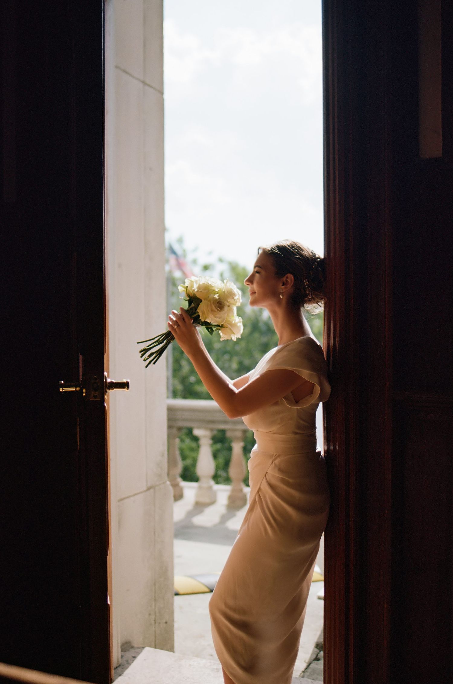 A silhouette captures an elegant bride in a form-fitting dress admiring her bouquet by a doorway.
