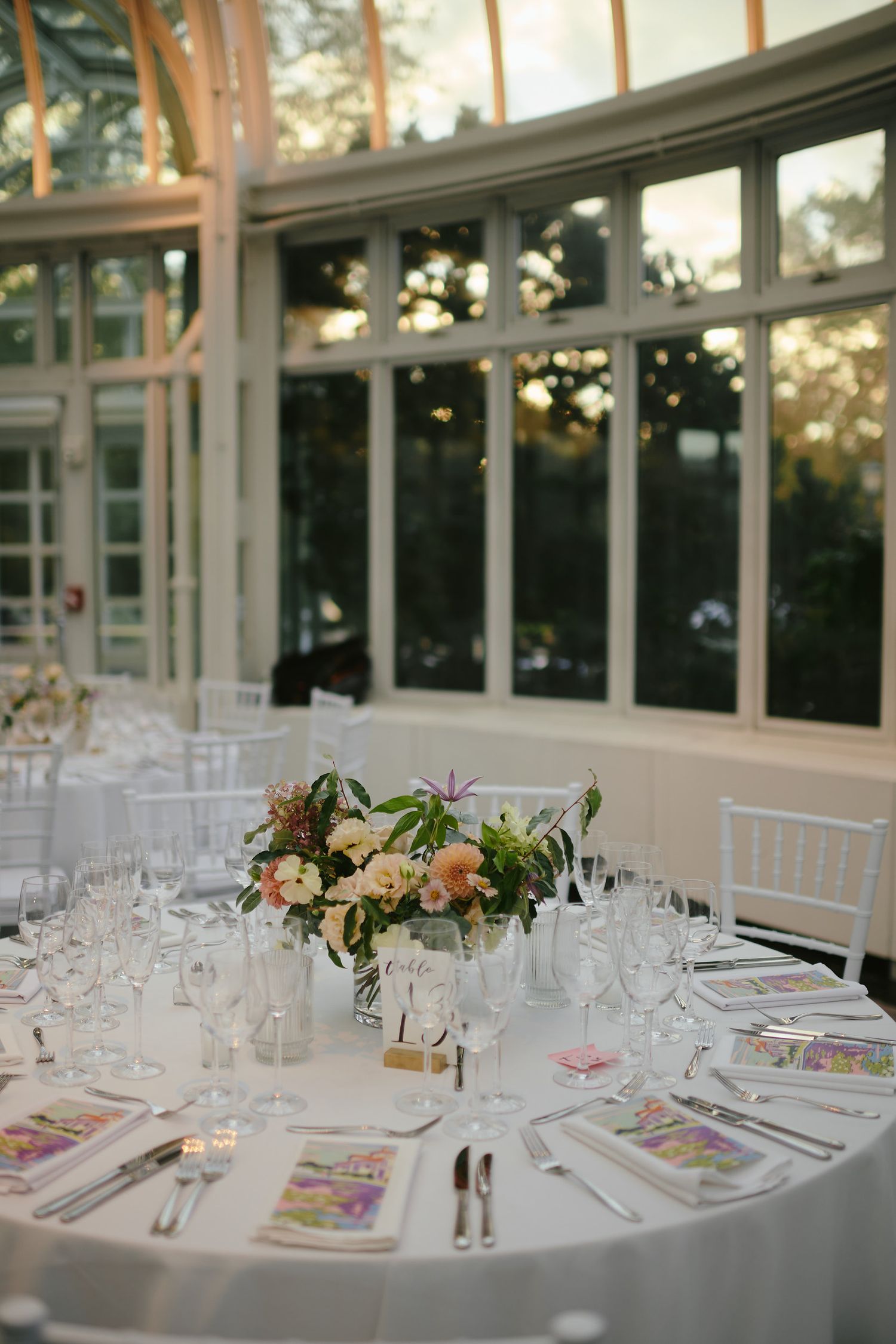 Elegant wedding reception table setting with floral centerpiece and wine glasses against floor-to-ceiling windows at sunset.