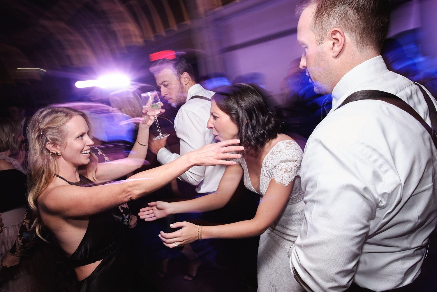 Guests dance energetically on a crowded dance floor at a wedding reception with purple and blue lighting effects.