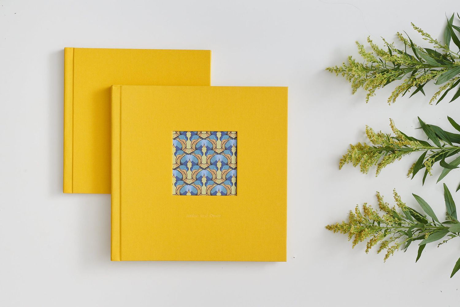 Two yellow square photo albums with decorative pattern inlay displayed next to green pine branches on white surface.
