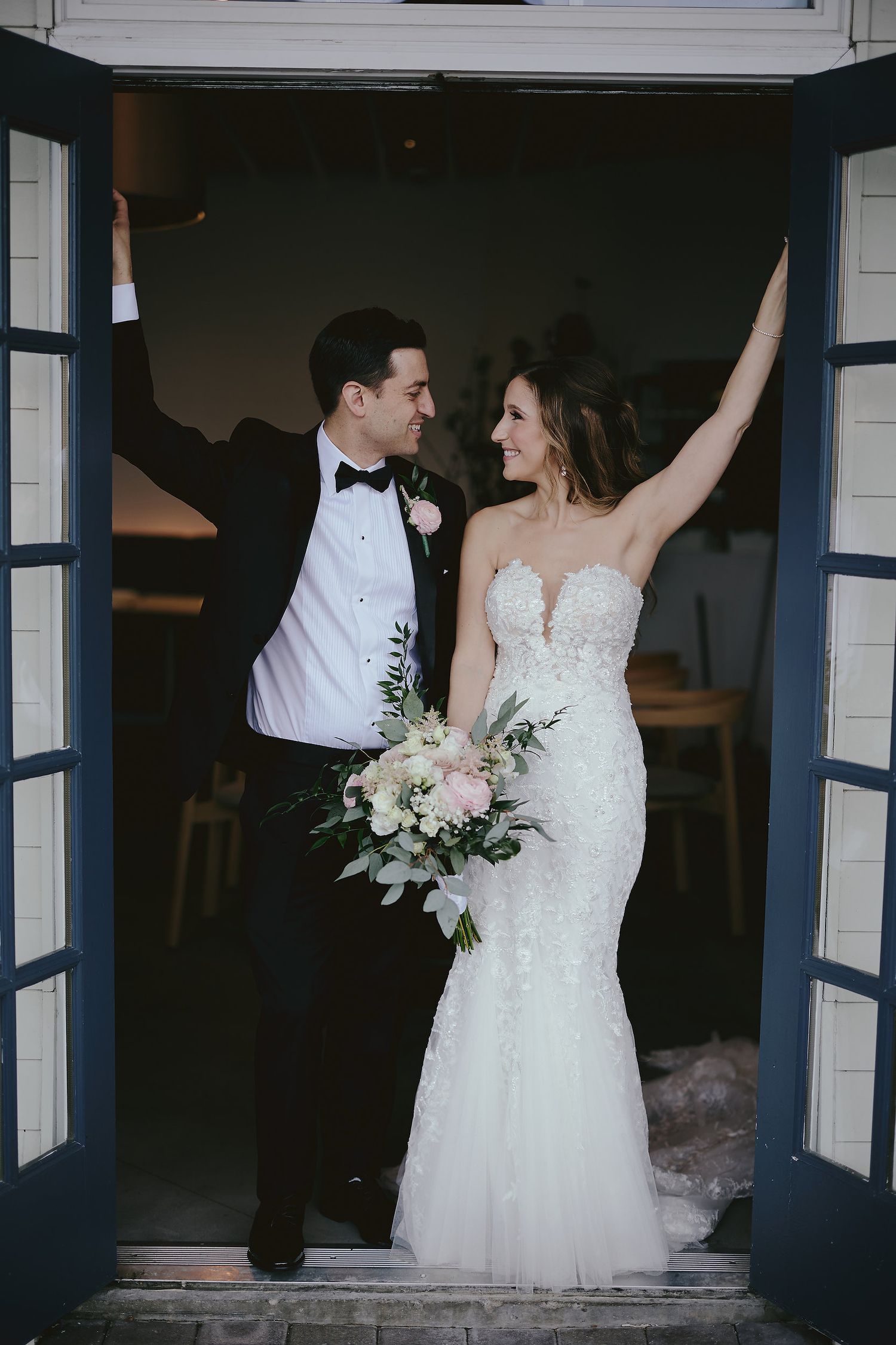 Newlyweds pose joyfully in white dress and tuxedo while framed by open blue doors at their wedding celebration.