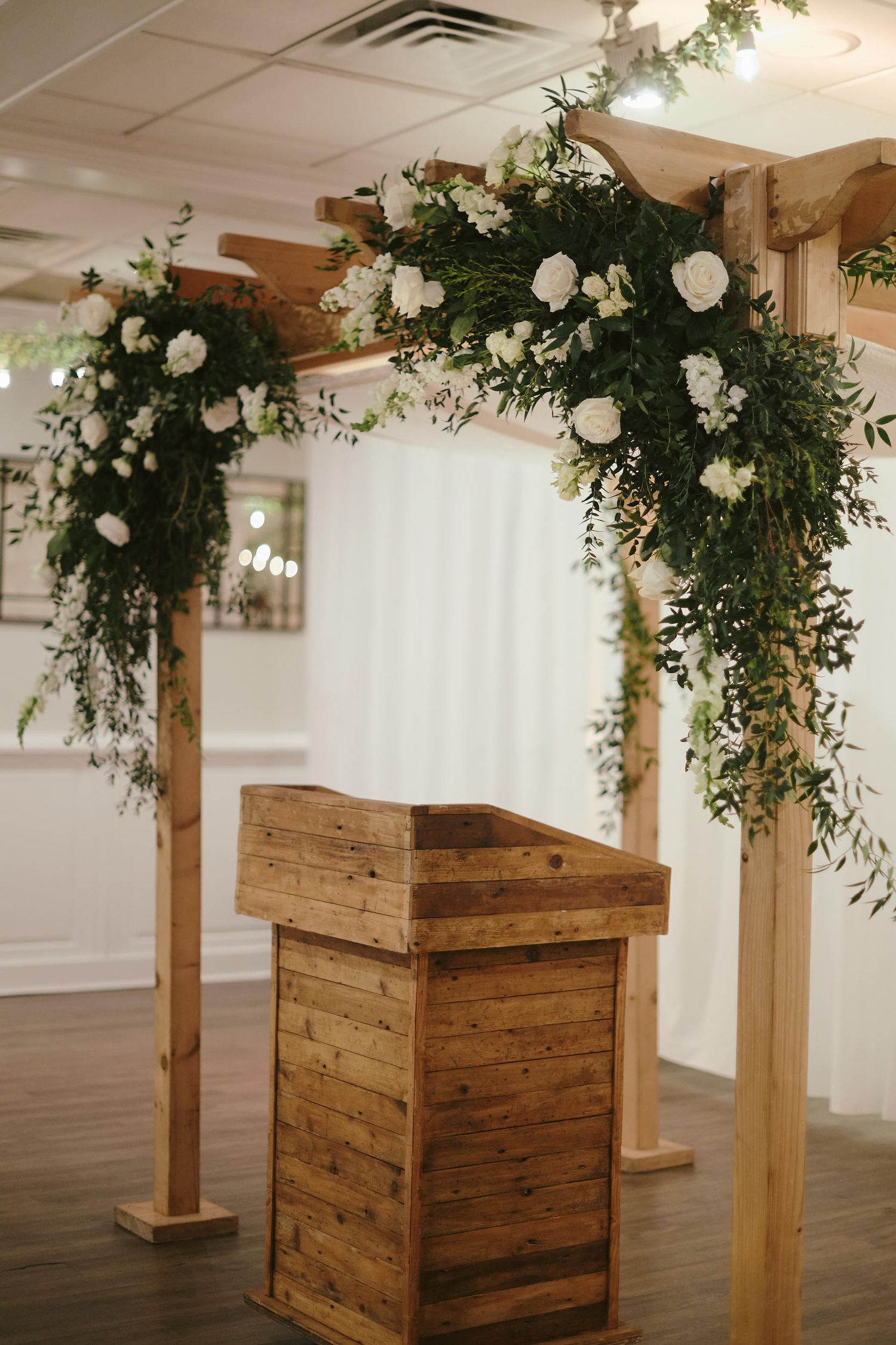 A wooden wedding arch decorated with lush greenery and white roses stands above a rustic podium in an indoor venue.