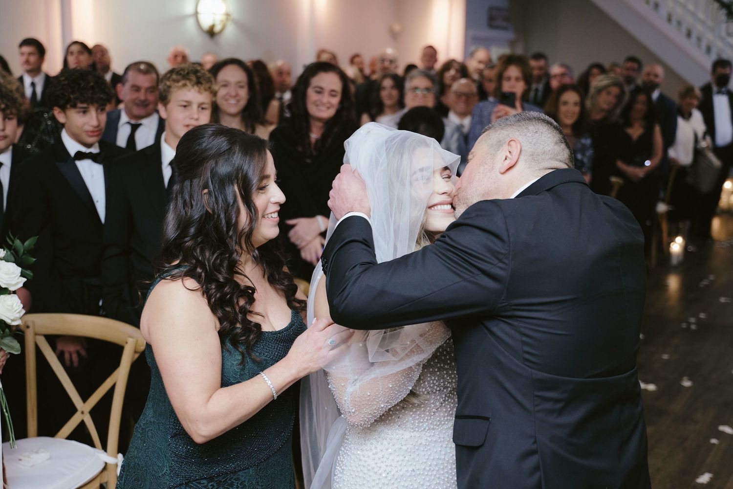 Three people share an emotional moment with embraces during an indoor formal event with guests in background.