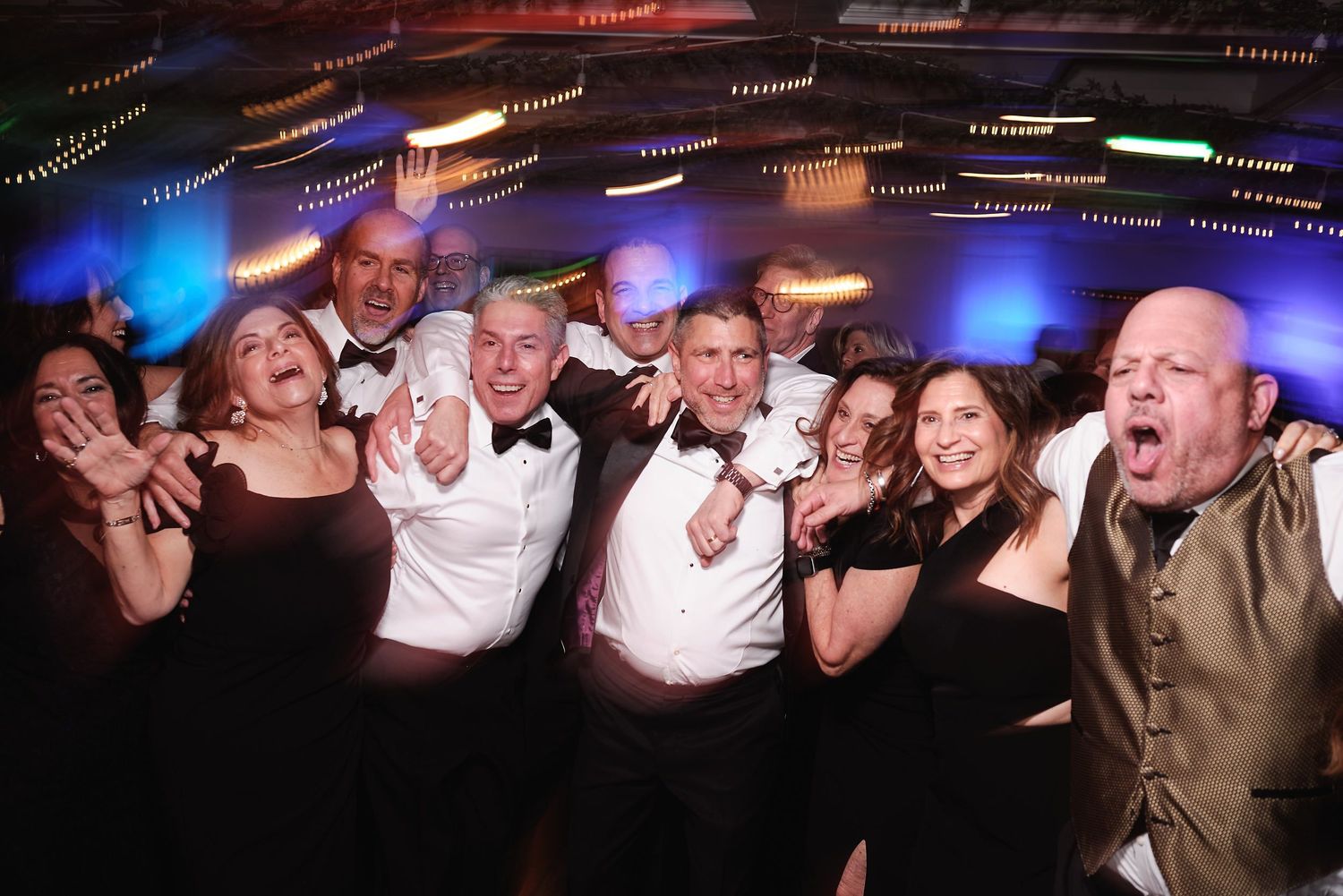 A lively group of party guests pose together under colorful ceiling lights at a celebration event.