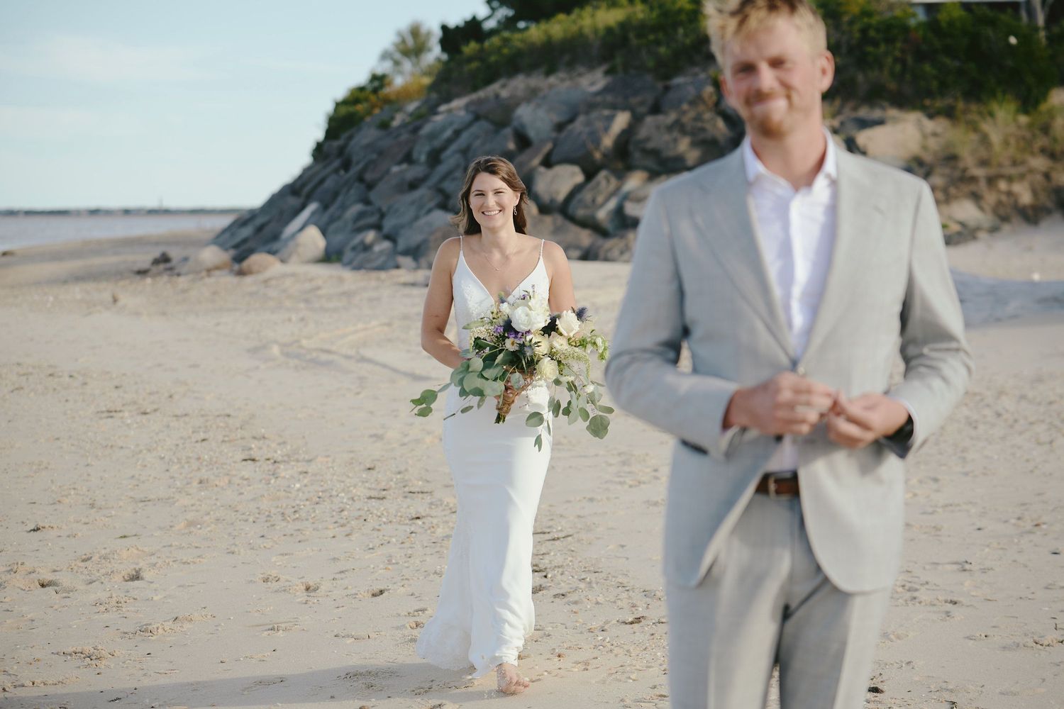 A couple in elegant wedding attire walk along a scenic beach with rocky cliffs in the background during sunset.